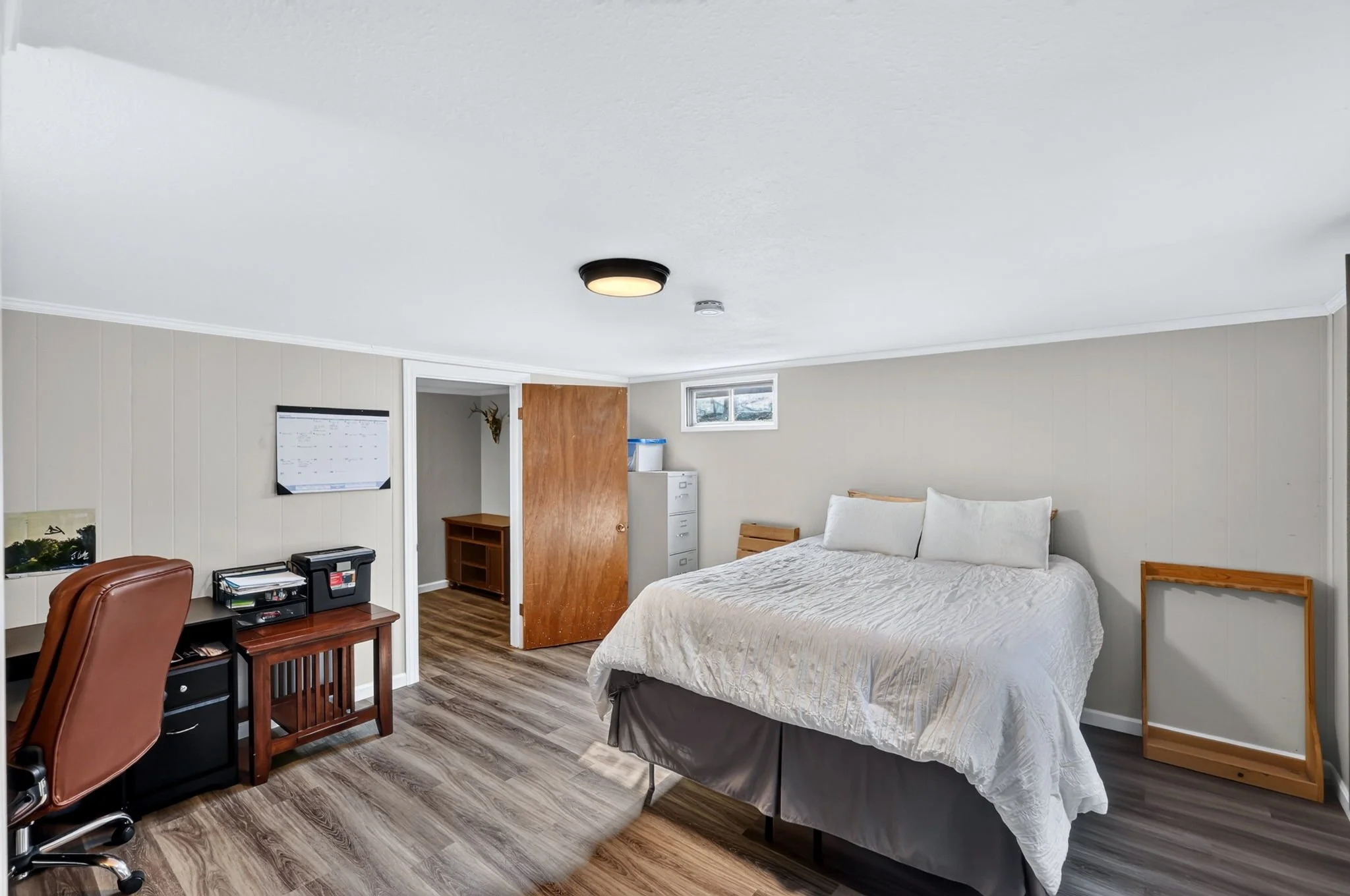 A bedroom with a bed, a wooden chair, a desk, and a small white cabinet near the wall, with beige paneled walls and wooden flooring.