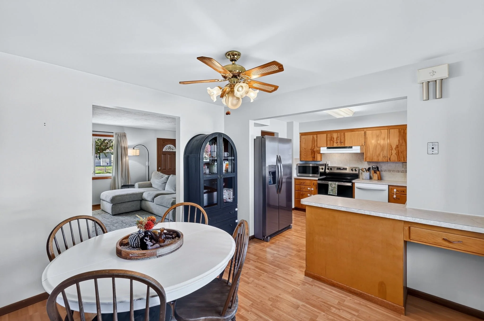 Interior of a kitchen and living room with a white ceiling fan, wooden cabinetry, a stainless steel refrigerator, and a white dining table with chairs.