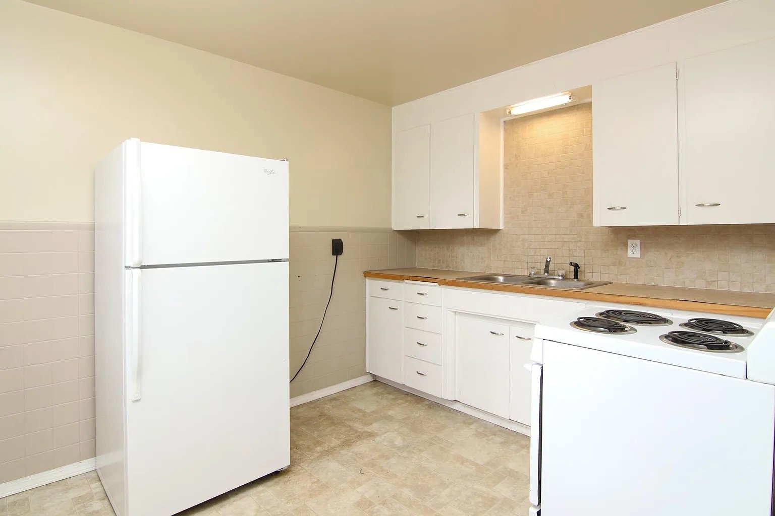 A simple kitchen with white cabinets, a white electric stove, a white refrigerator, a double sink, beige tiled backsplash, and beige linoleum flooring.