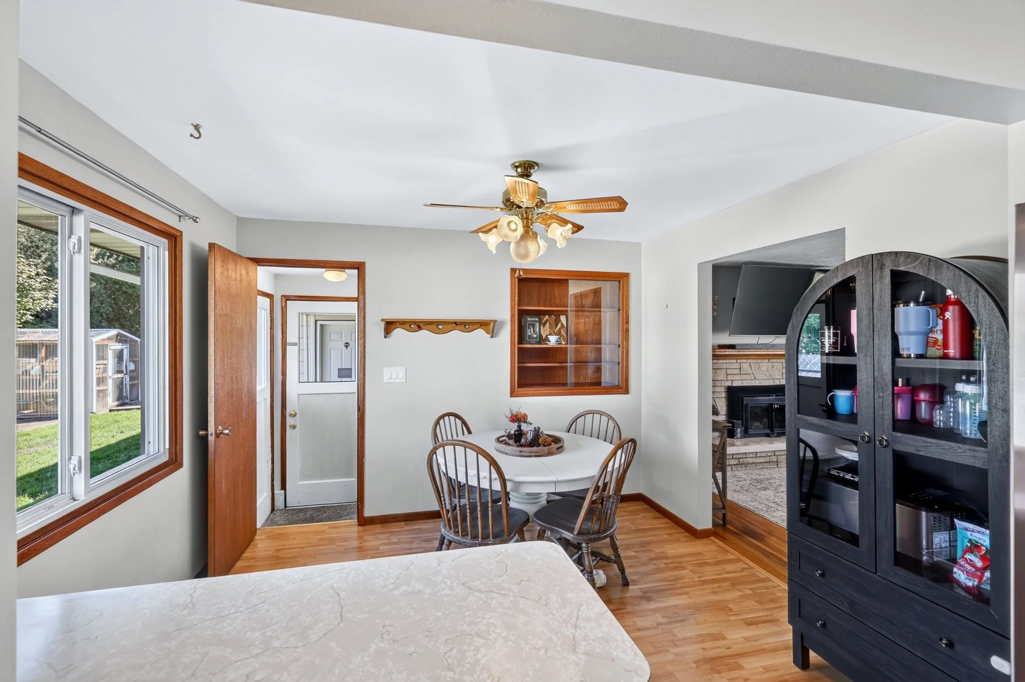 The image depicts a cozy dining area inside a home, with a round wooden table and four chairs. There's a window with wooden trim and a sliding glass door leading to the outside. A ceiling fan with light fixtures hangs from the ceiling. The room has a