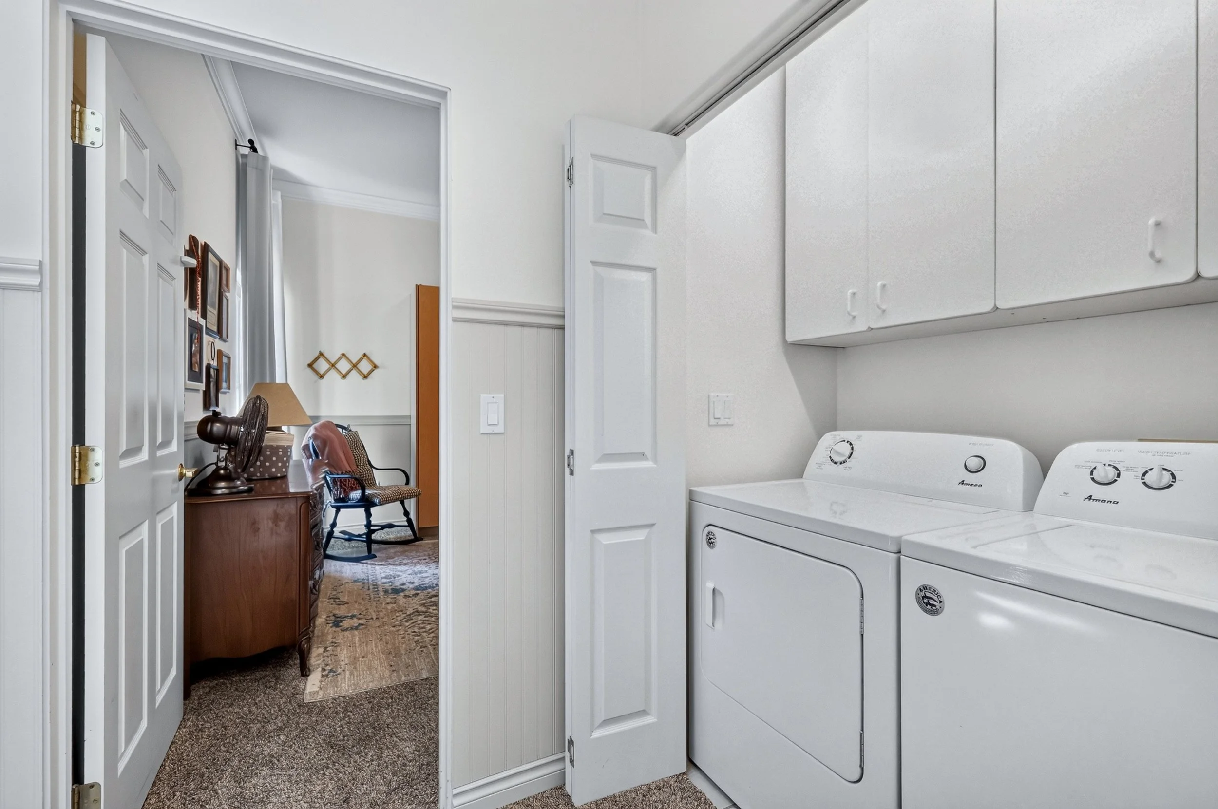 A laundry room with white washer and dryer and white cabinets, with a doorway leading to a living area with a table, chair, and wall decorations.