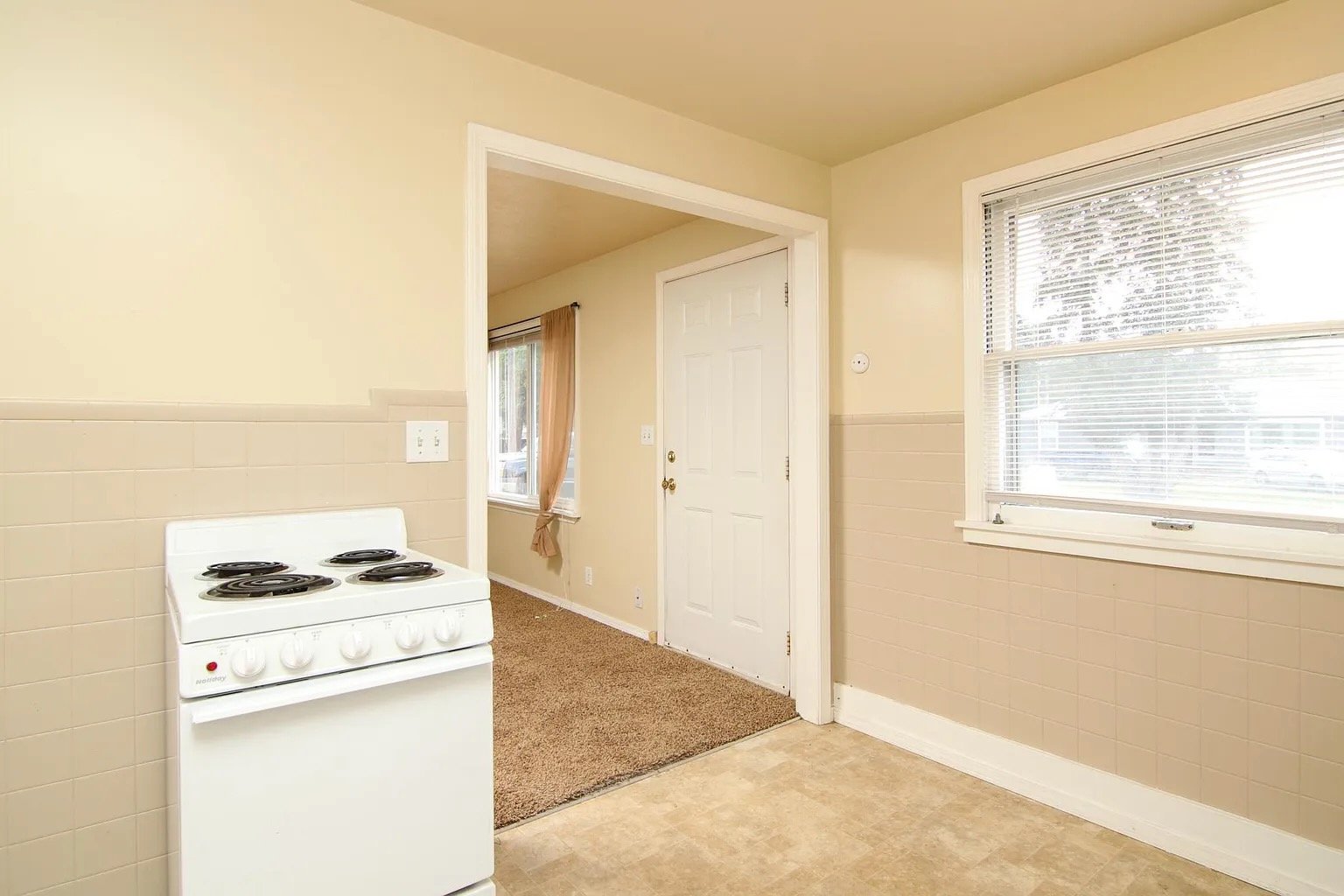 Kitchen area with a white electric stove, beige tiled walls, a window with blinds, and a doorway leading to a carpeted room with a window and curtains.