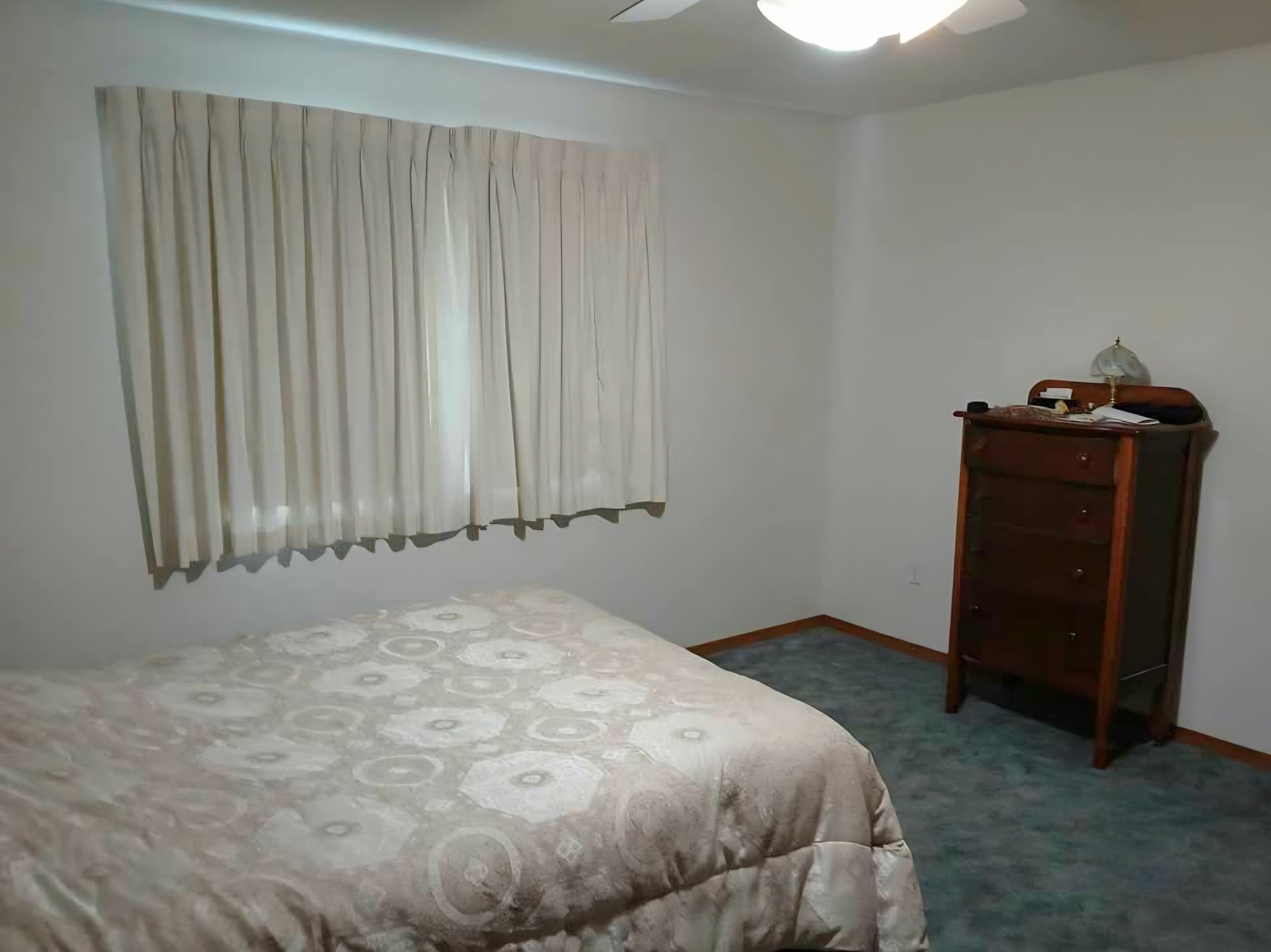 A bedroom with white walls and a window covered by cream-colored curtains, a patterned bedspread on the bed, a small wooden dresser with a lamp and some objects on top, and carpeting on the floor.