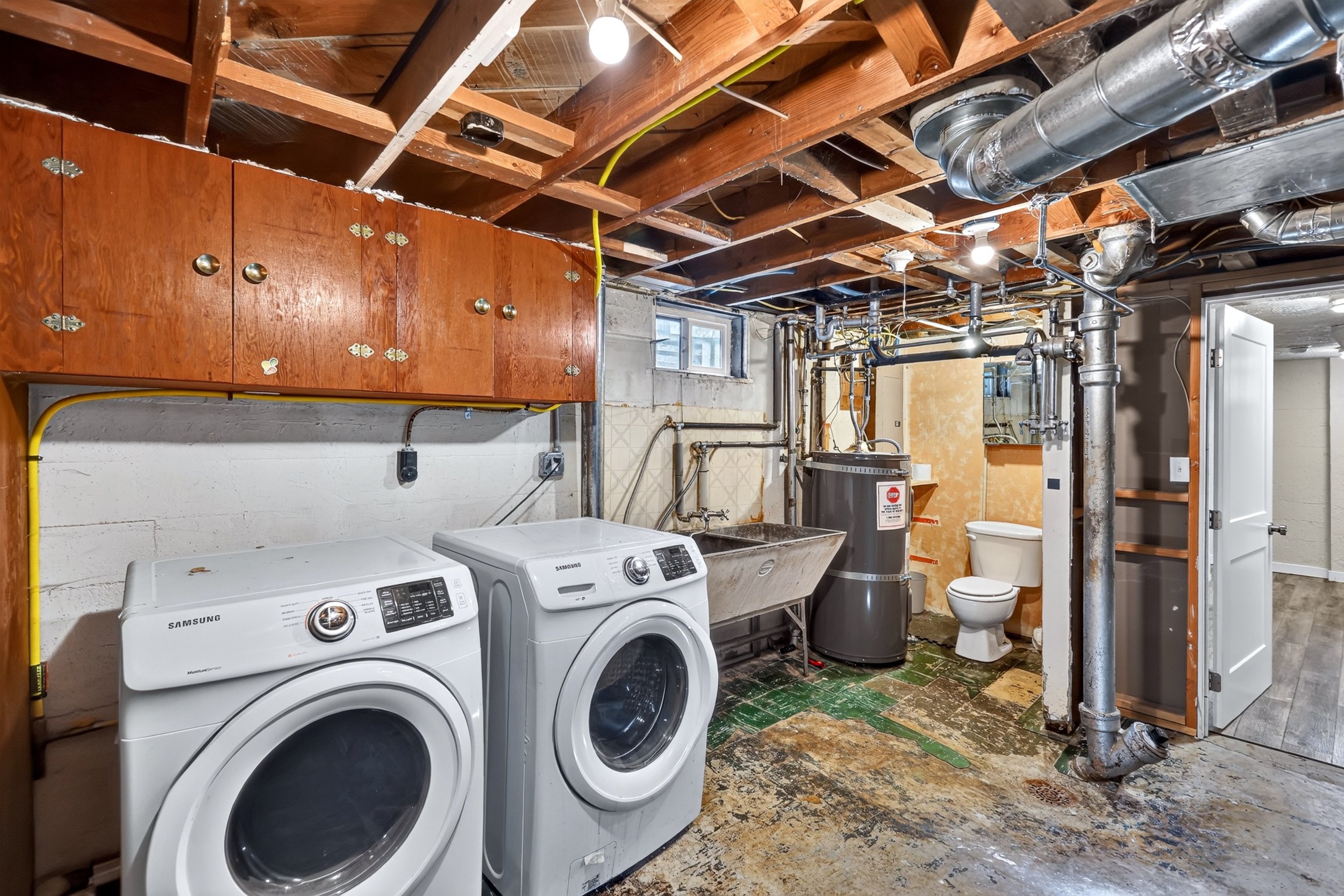 A laundry room under construction with exposed wooden ceiling beams, a small window, two front-loading Samsung washers, a utility sink, a toilet, and exposed ductwork and pipes.