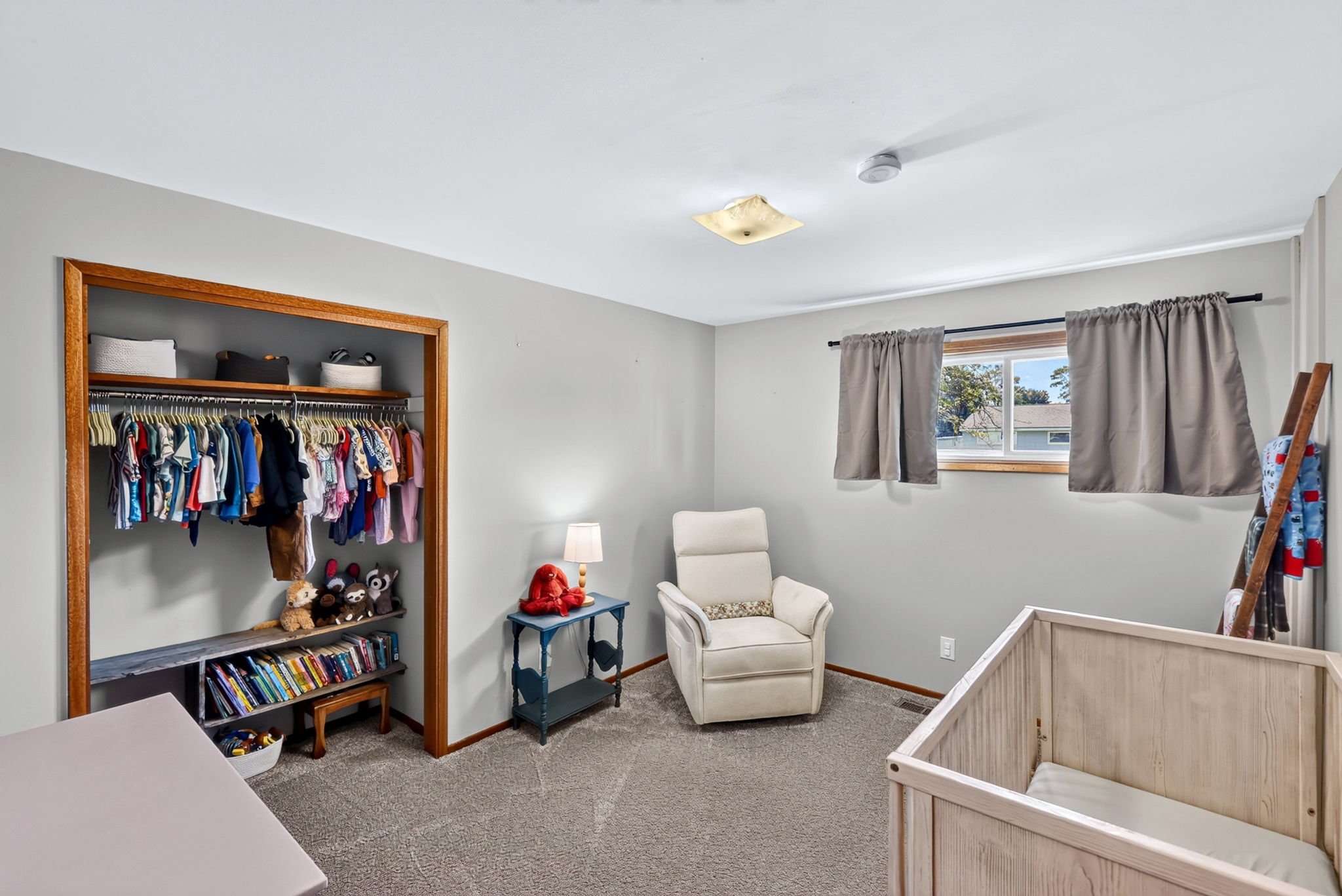A cozy bedroom with a white armchair, a small blue side table with a red stuffed animal, a wooden bookshelf filled with books and toys, and a white crib in the corner. There is a window with beige curtains letting in natural light, a ceiling light fi
