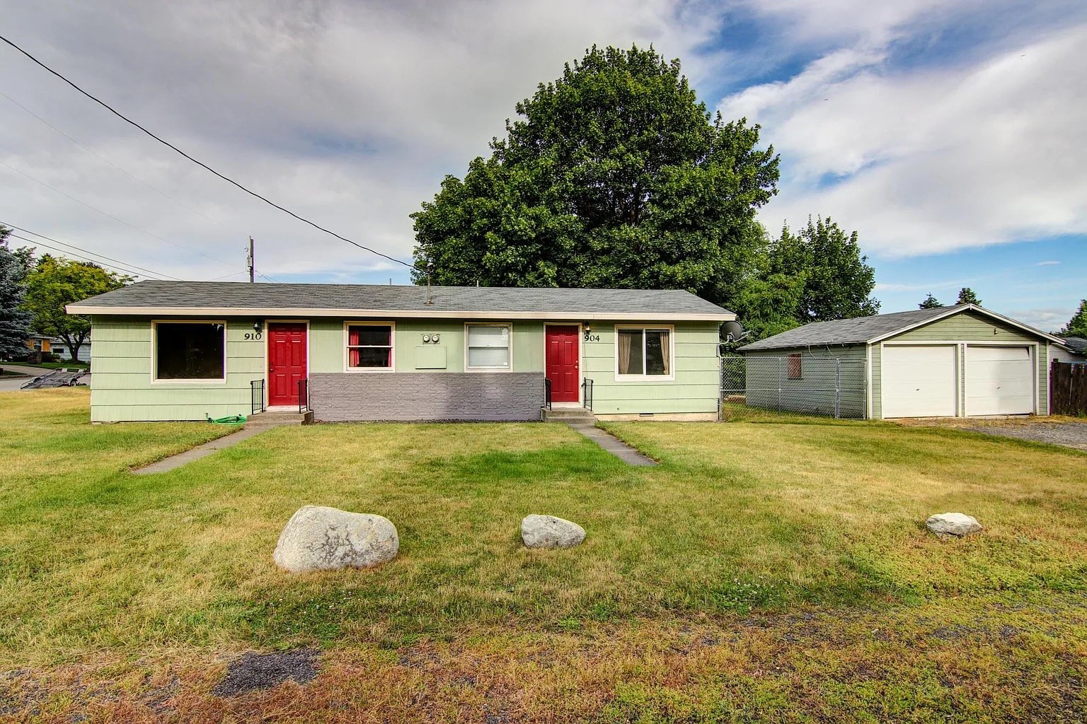 A small, light green house with red doors, a front window with pink curtains, and a gray lower facade. There are two concrete walkways leading to the doors, with three large rocks in the front yard. Behind the house is a large tree, and to the right 