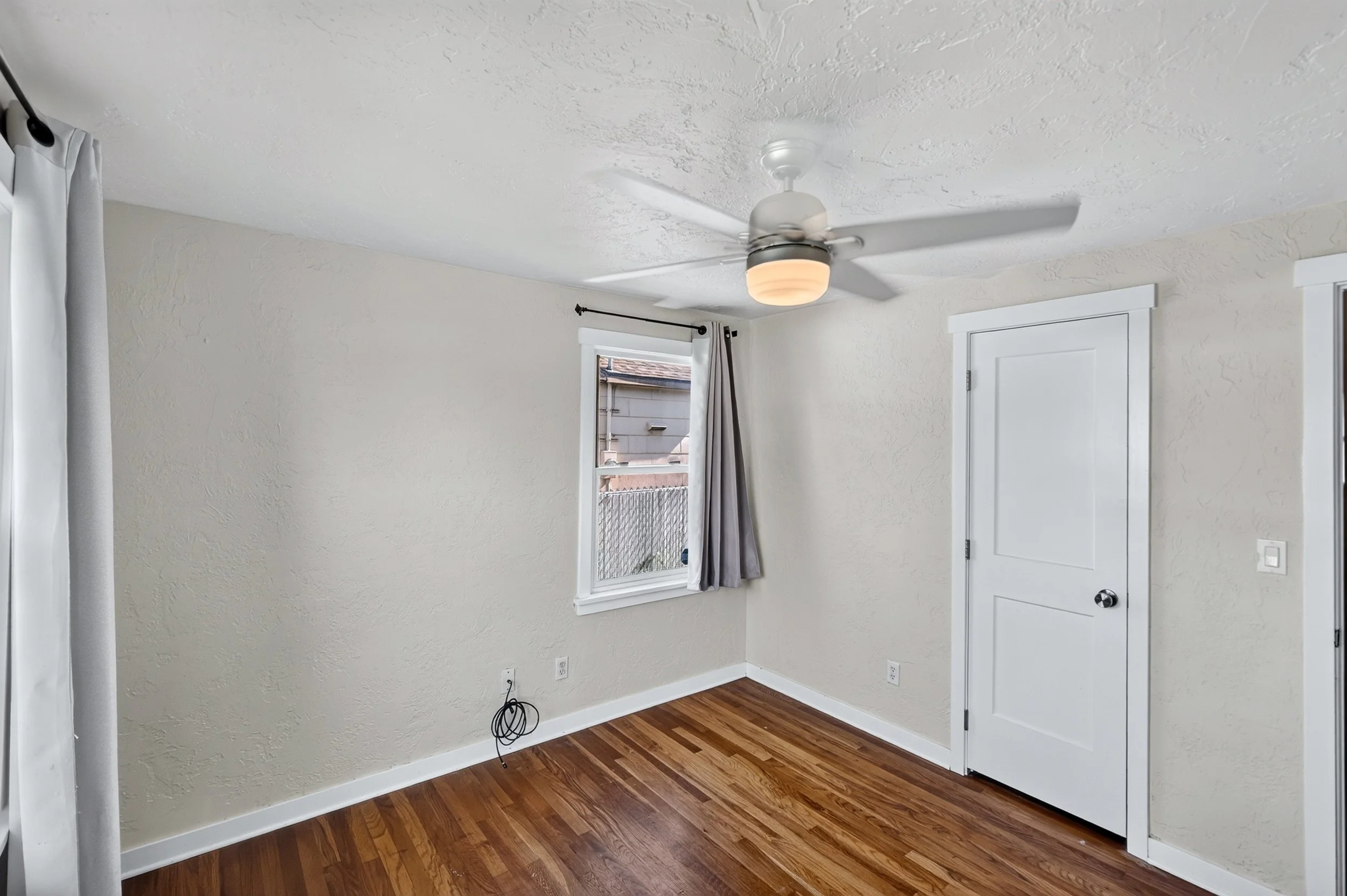 Interior view of a room with white walls, hardwood flooring, a ceiling fan with a light, a window with curtains, and a door.