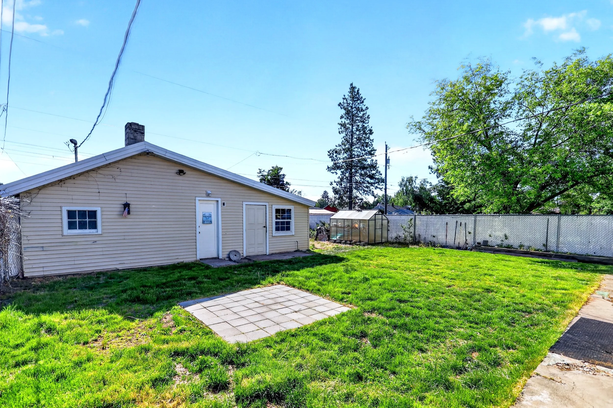 A backyard with green grass, a small paved area, a house with yellow siding, a white door, a window, a fence, tall trees, a greenhouse, and a clear blue sky.