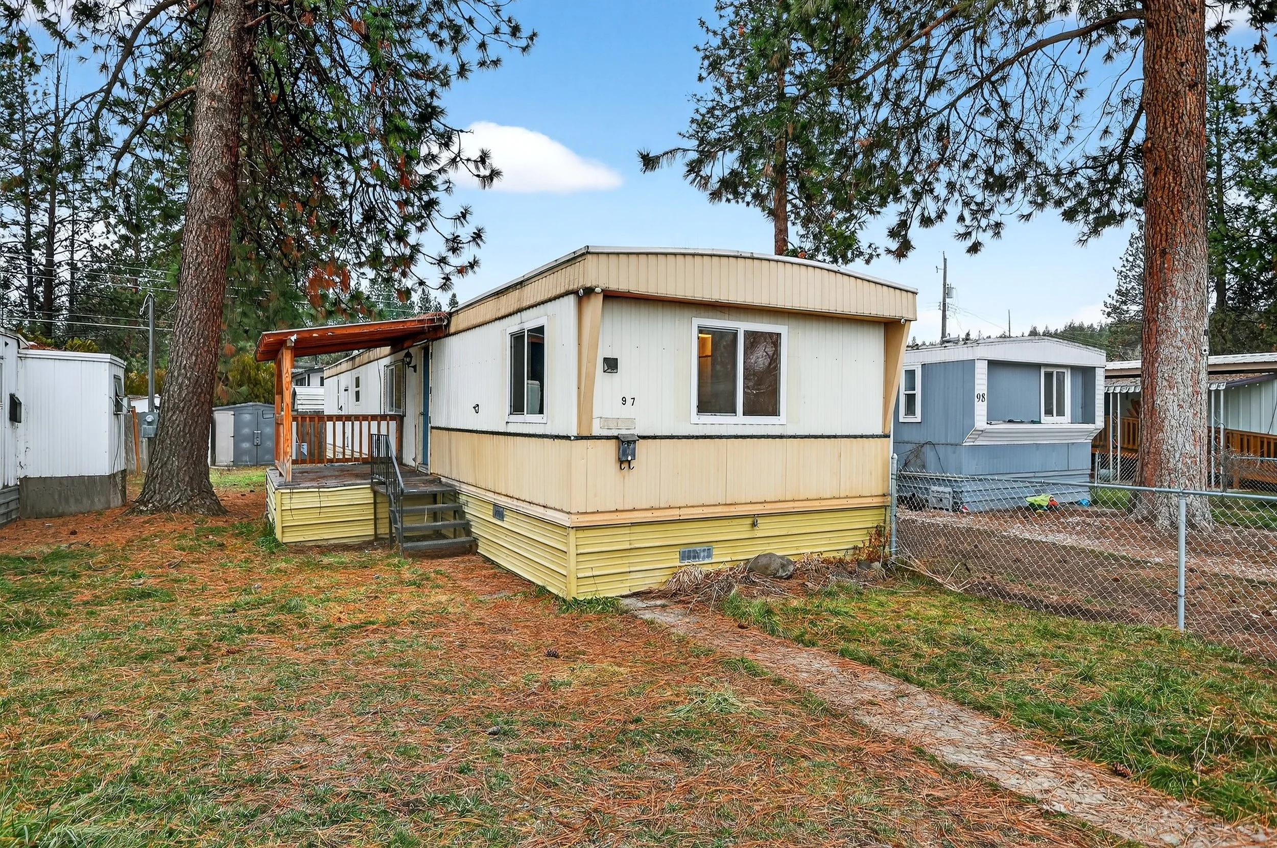 A mobile home with beige and light blue exterior, located in a wooded area with large pine trees, trees, a chain-link fence, and a yard with fallen leaves and pine needles.