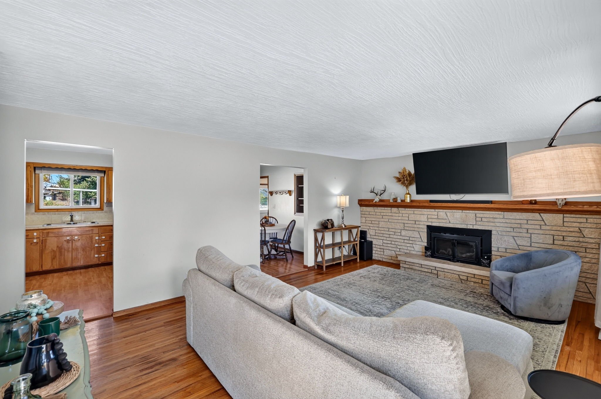 Living room with a beige sofa, a gray armchair, a brick fireplace with a large TV mounted above, and a wooden mantel decorated with a vase and a deer head sculpture. Adjacent to the living room, a dining area and kitchen are visible, with wooden floo