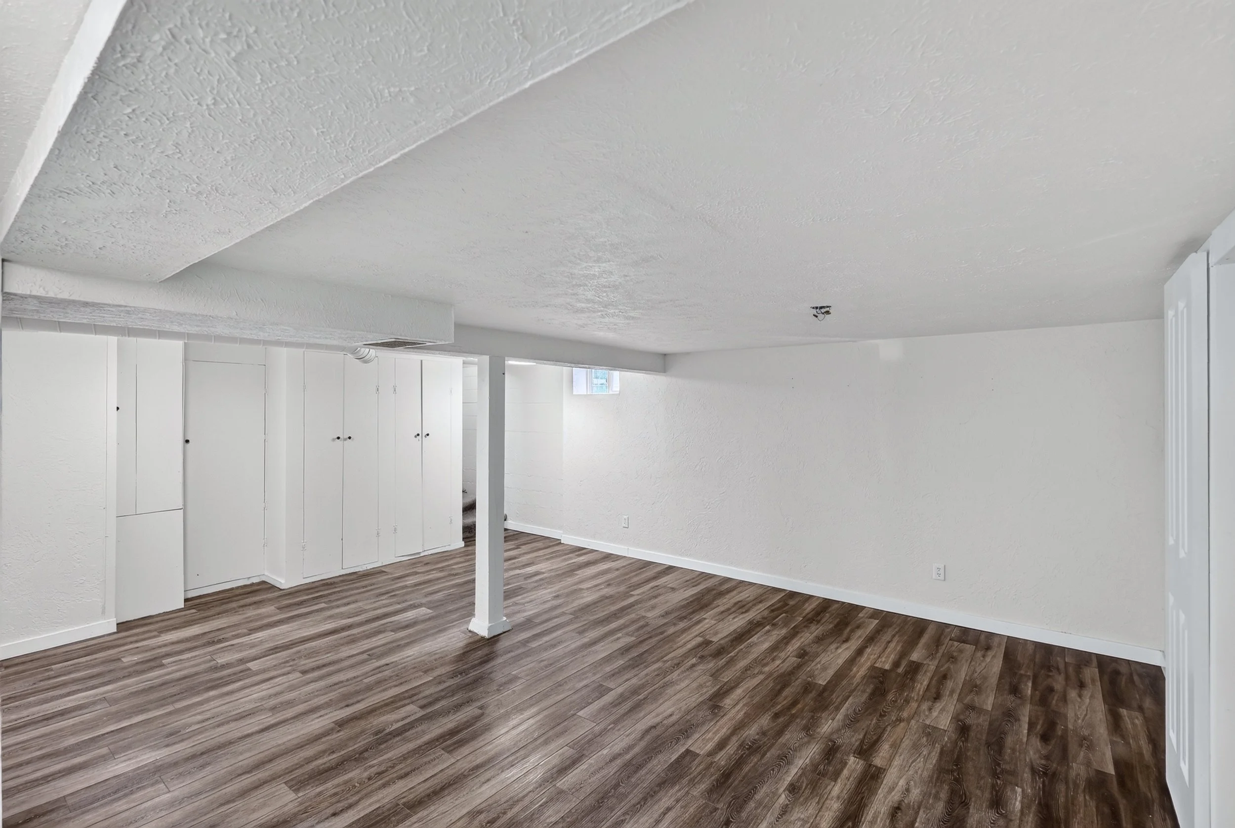 Empty room with white textured walls, wood laminate flooring, and a white ceiling with a light fixture. There are white cabinets along one wall and a small window letting in natural light.