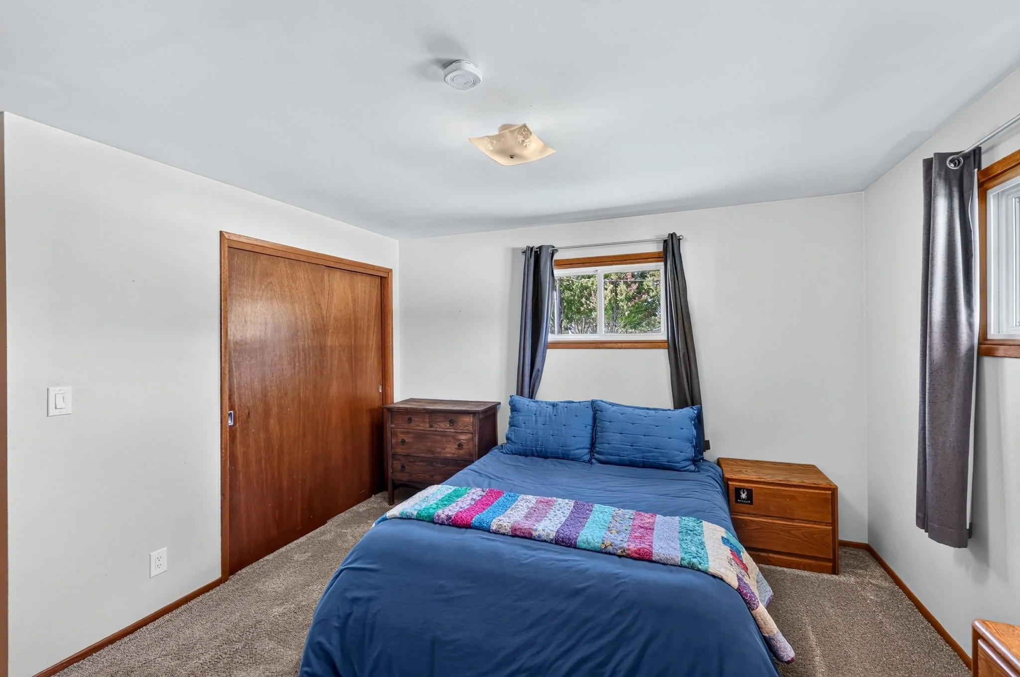 A bedroom with white walls, a brown wooden door, a small wooden dresser, a window with gray curtains, a bed with a colorful quilt, and a ceiling light fixture.