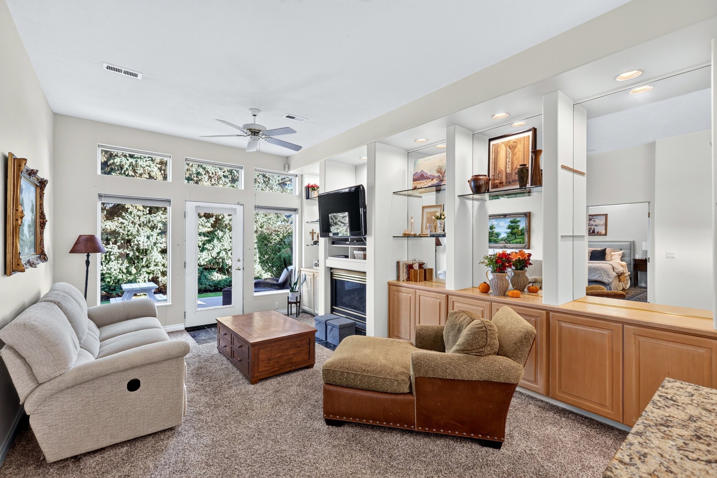 Living room with beige sofa, armchair, wooden coffee table, large windows, a ceiling fan, and a view of outdoor greenery.