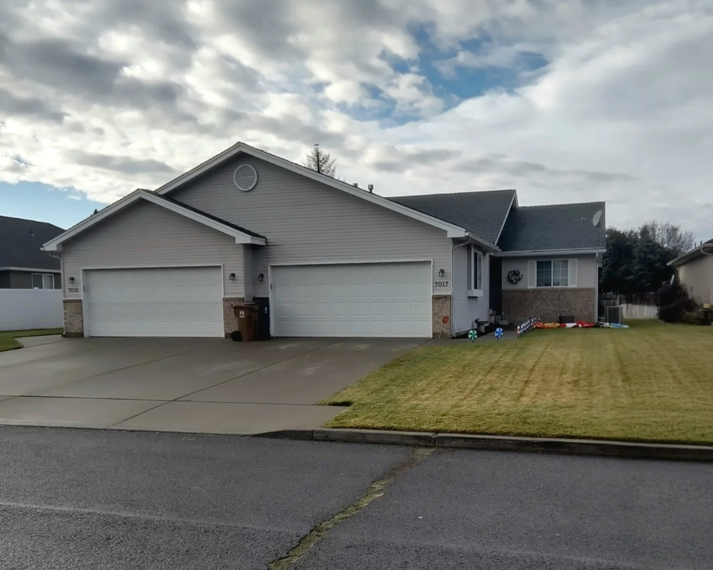 A suburban house with a double garage, gray siding, and brick accents, front lawn, and a driveway with a trash bin, under a partly cloudy sky.