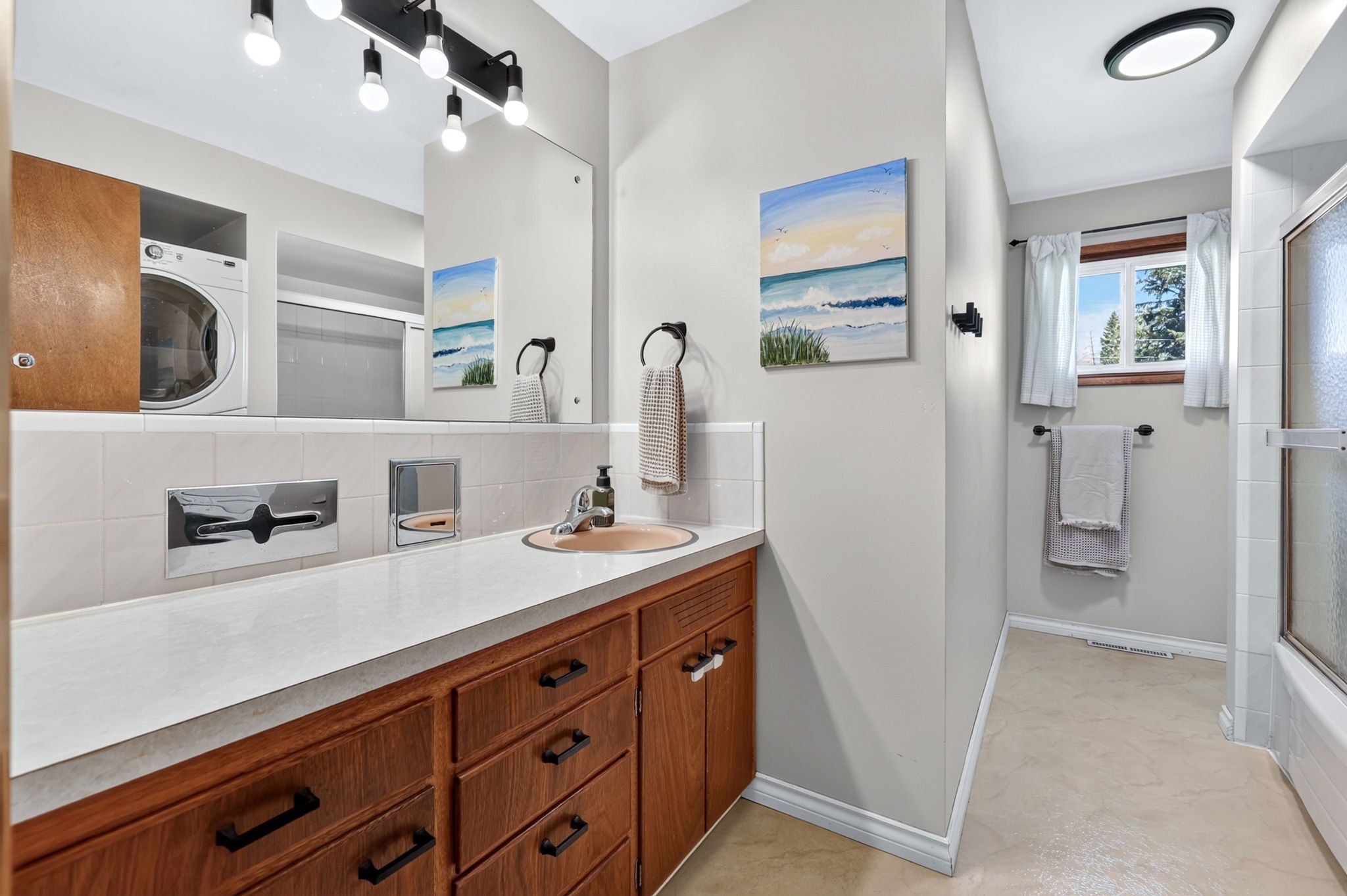 A bathroom with a wood vanity, beige countertop, pink sink, and a large mirror. The wall has two wall art paintings of the beach and ocean. To the right, there are white curtains on a window, a towel rail, and a walk-in shower with textured glass.