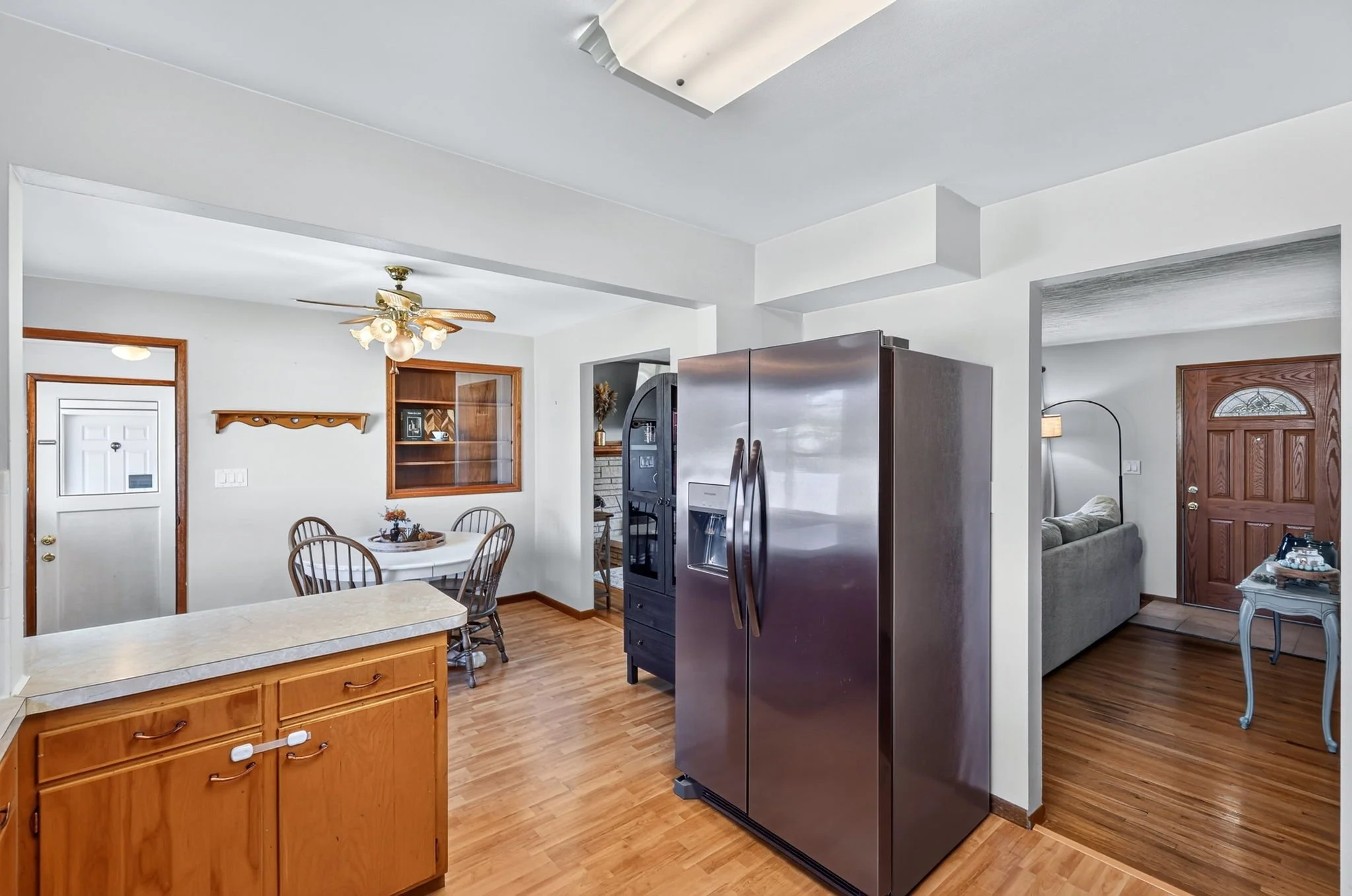Interior of a home kitchen and living area with a stainless steel refrigerator, dining table and chairs, wooden cabinets, a ceiling fan, and a living room with a gray couch and a wooden door.