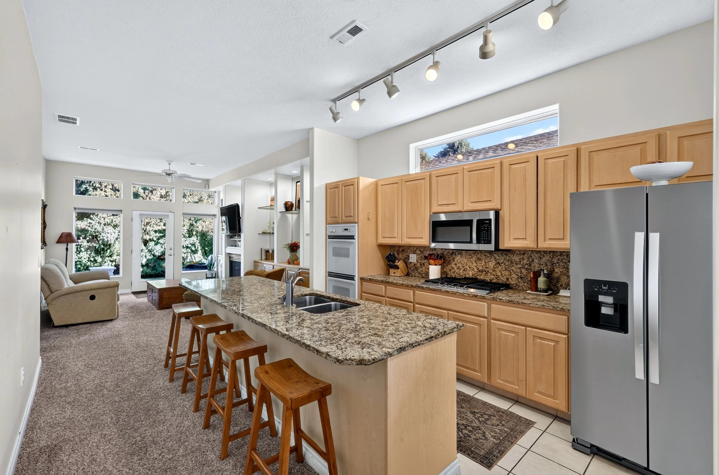 Open-concept kitchen and living room with large windows, wooden kitchen cabinets, granite countertops, stainless steel appliances, and beige carpeted seating area.