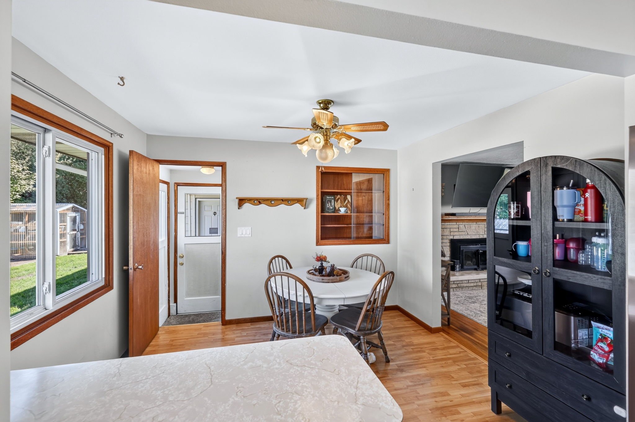 A cozy dining room with a round table, four chairs, a window showing the outdoors, a ceiling fan with light fixtures, and a view of a living area with a fireplace and a shelf with items.