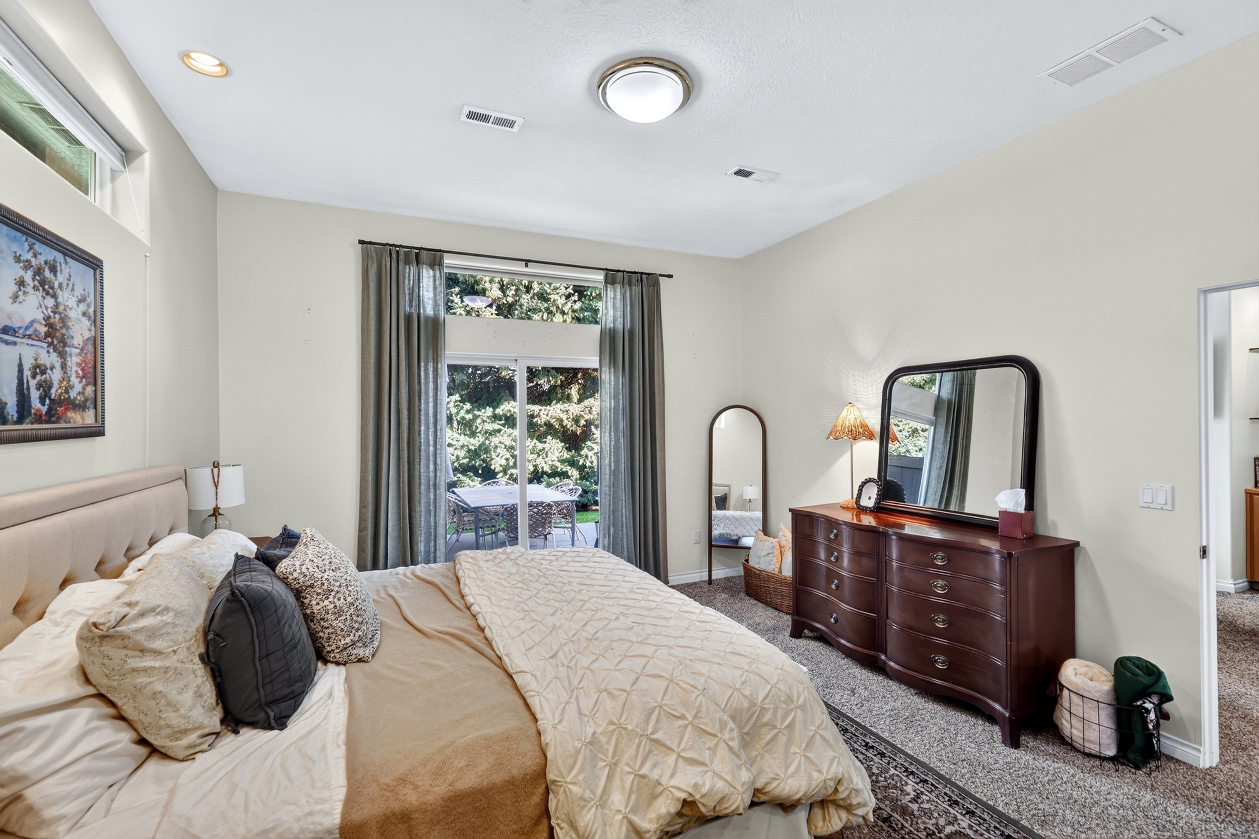 A cozy bedroom with a large bed covered in beige bedding and patterned pillows. There is a wooden dresser with a mirror and a small table lamp with a textured shade. A window with sheer curtains reveals an outdoor patio with chairs and trees. The cei