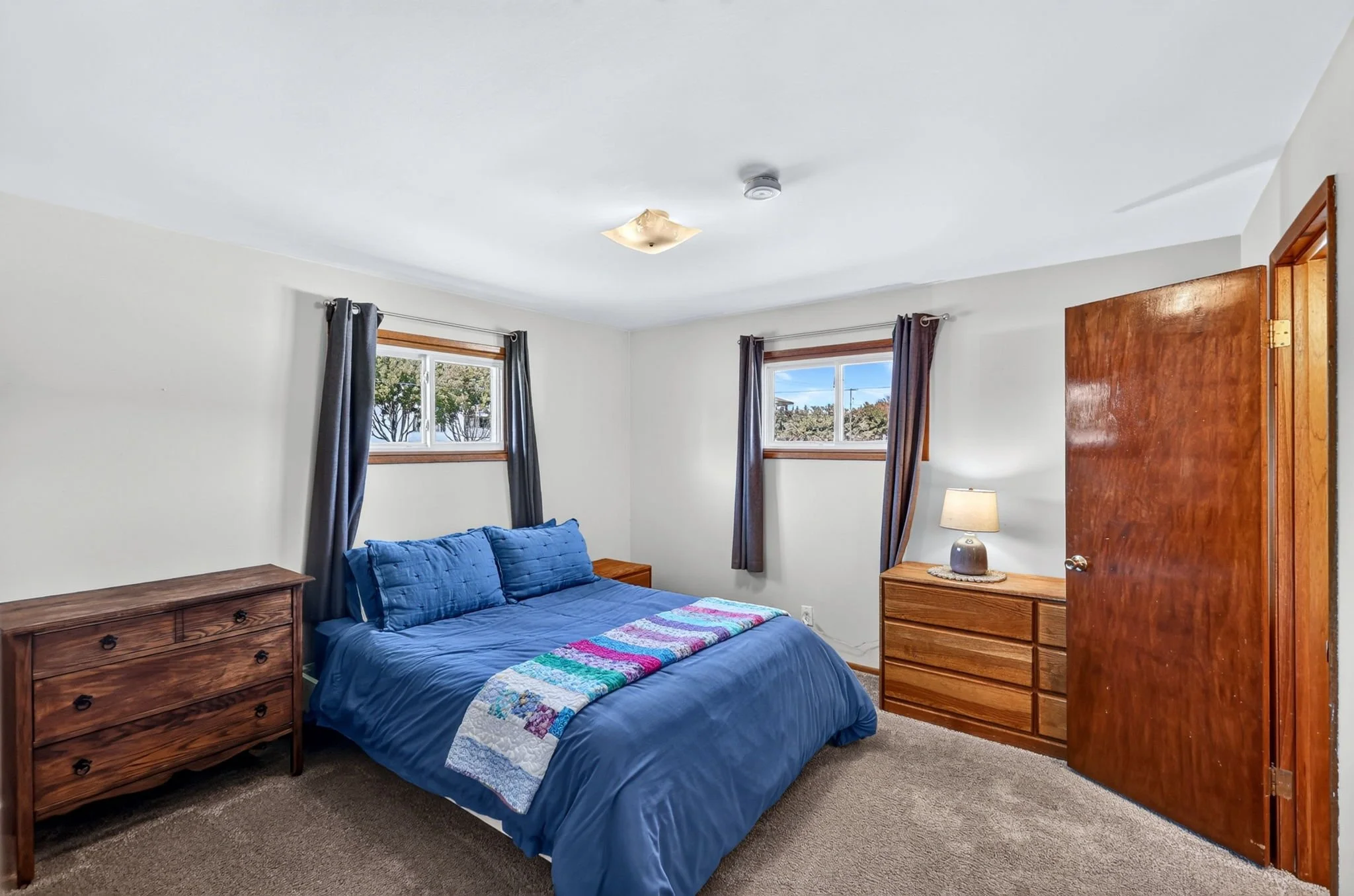 A cozy bedroom with a blue bedspread, wooden furniture, two windows with curtains, a small lamp, and a colorful quilt at the foot of the bed.