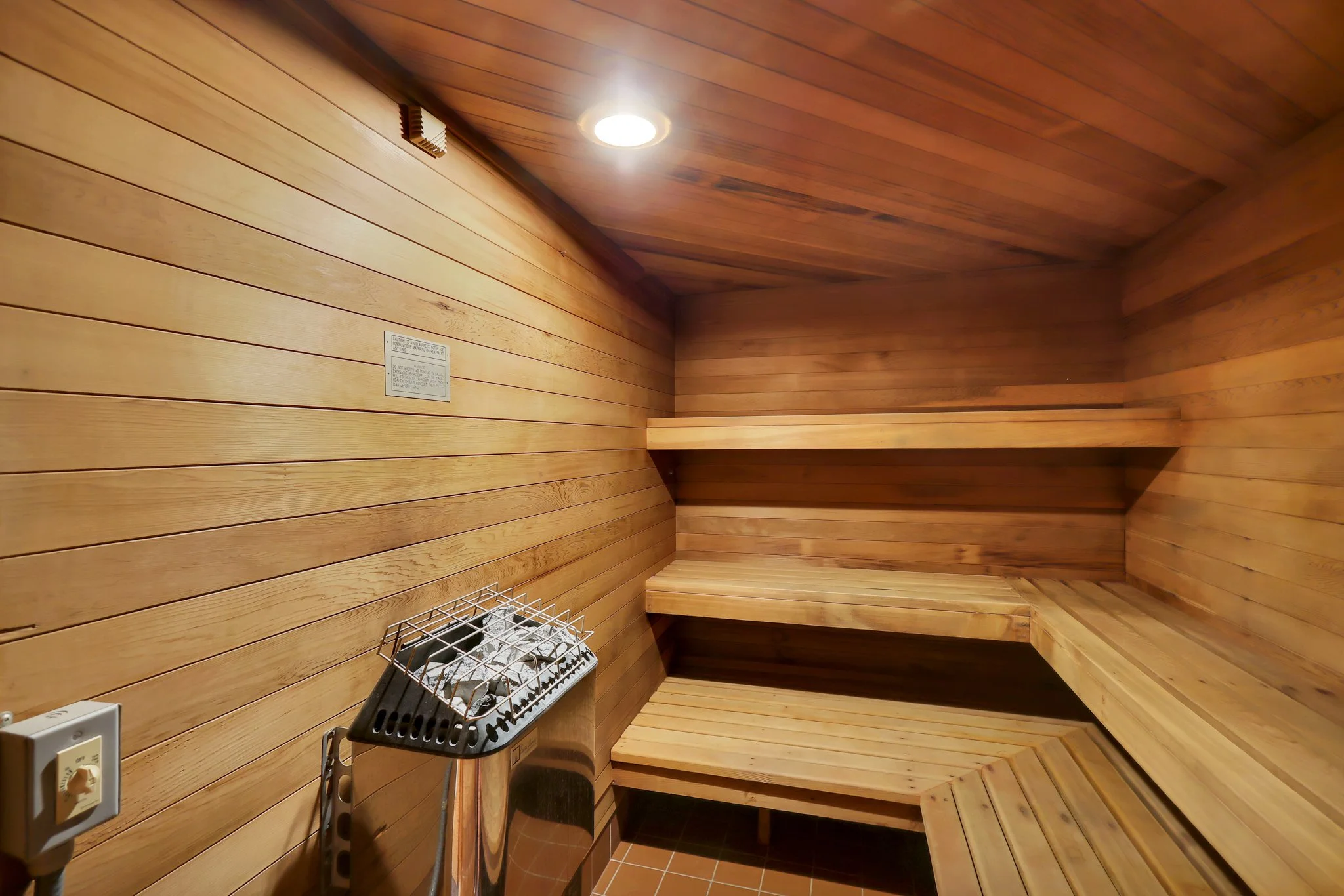 A wooden interior with horizontal wood paneling on the walls and ceiling, a light fixture on the ceiling, a metal heater, and a small bench.