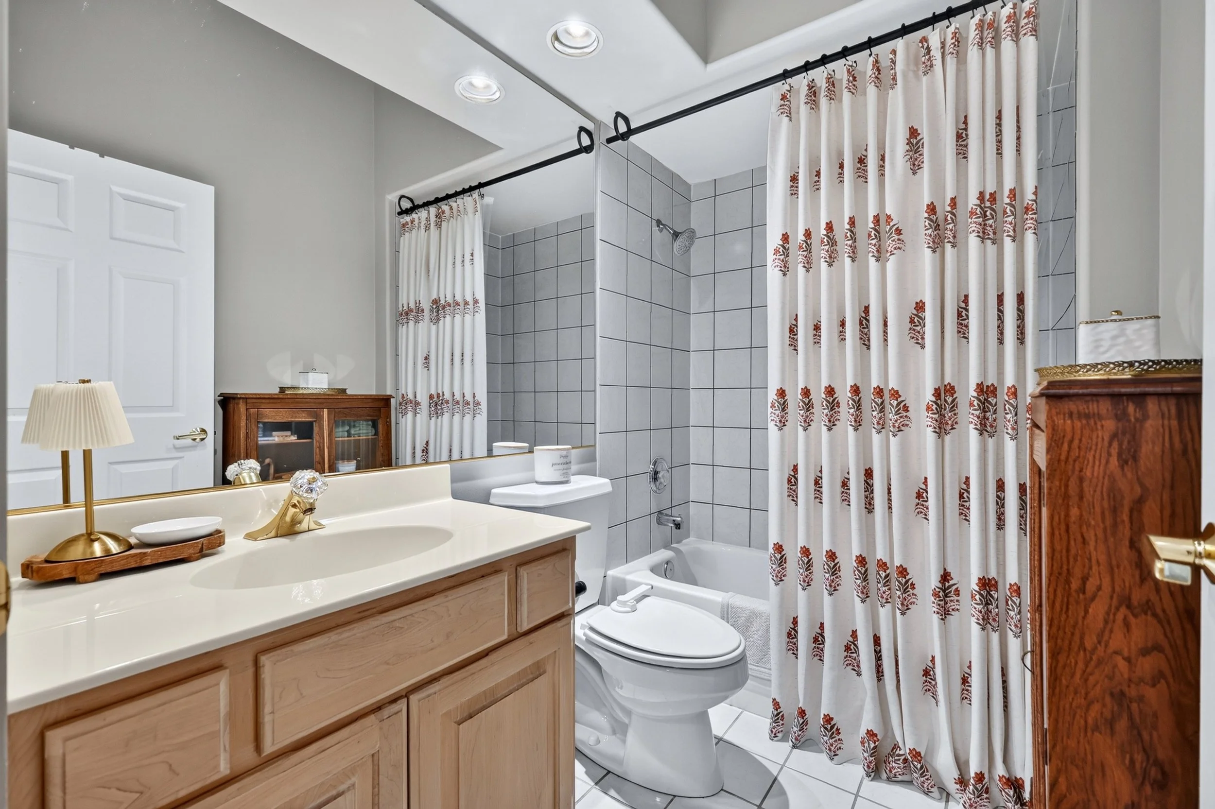 Bathroom with a white sink, a wooden cabinet, a toilet, a bathtub with gray tile walls, a showerhead, and a patterned shower curtain with red and brown designs.