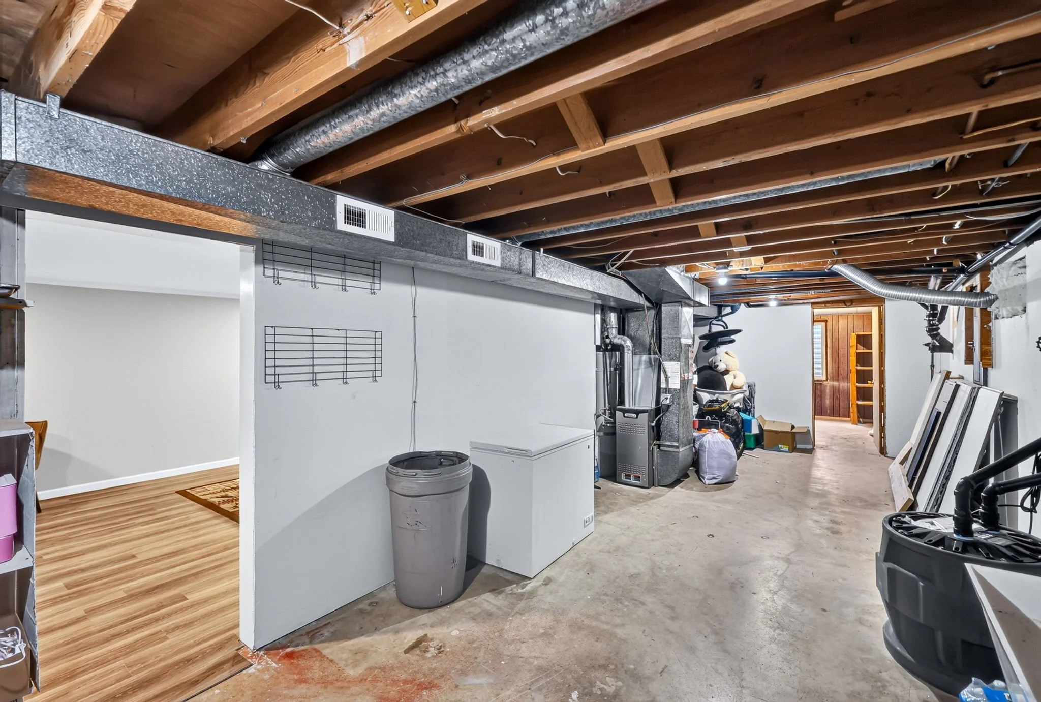 Unfinished basement with exposed wooden ceiling beams, ductwork, and ventilation pipes. There is a laundry area with a washing machine and dryer, and various boxes and items scattered around.
