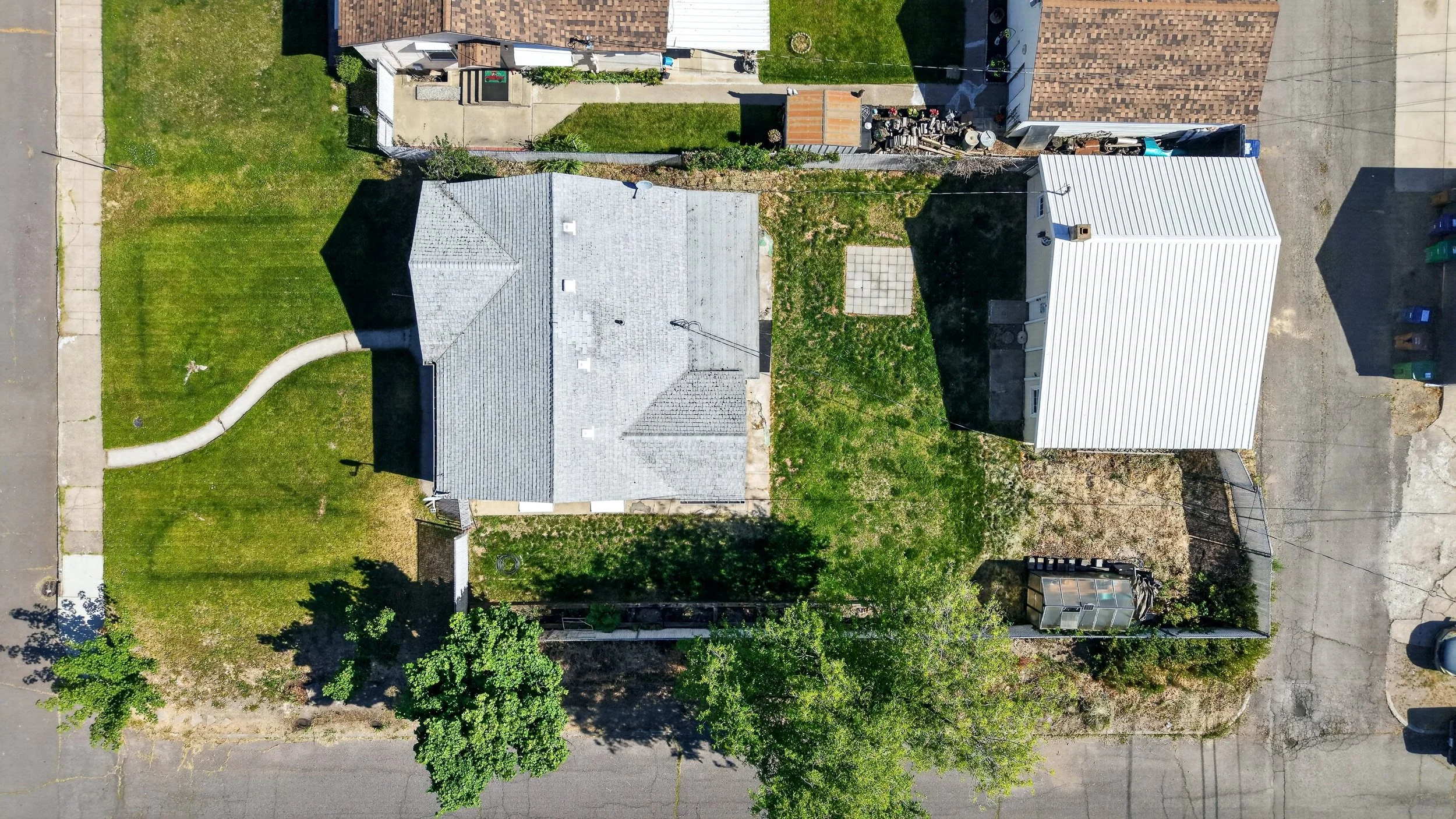 Aerial view of two houses with yards, one with a gray roof and the other with a white roof, separated by a narrow alleyway. The yards have grass, concrete walkways, and outdoor items. Streets are visible on the top and bottom edges.