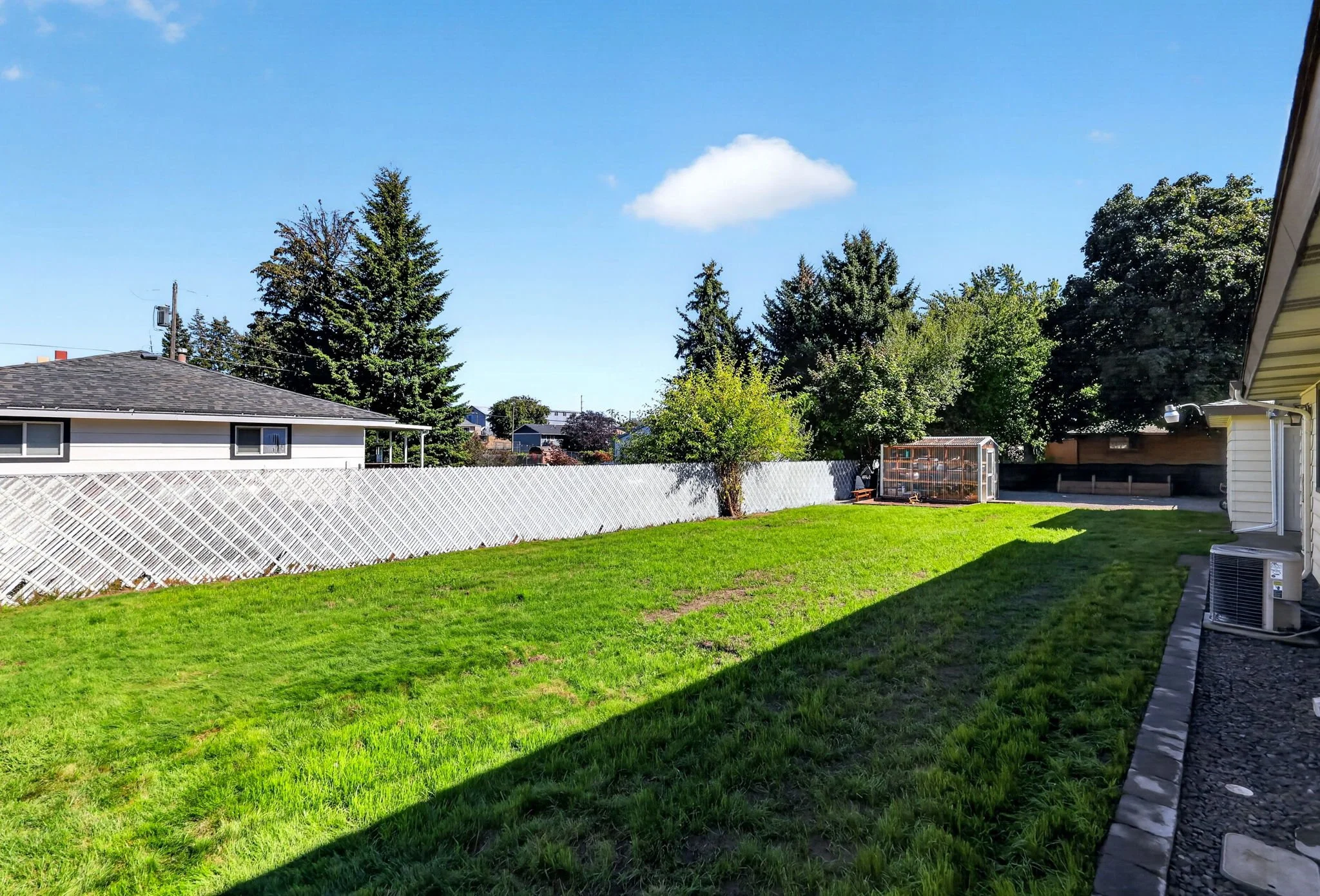 A backyard with a green lawn, trees, a white fence, a house with a gray roof, and a clear blue sky with a few clouds.