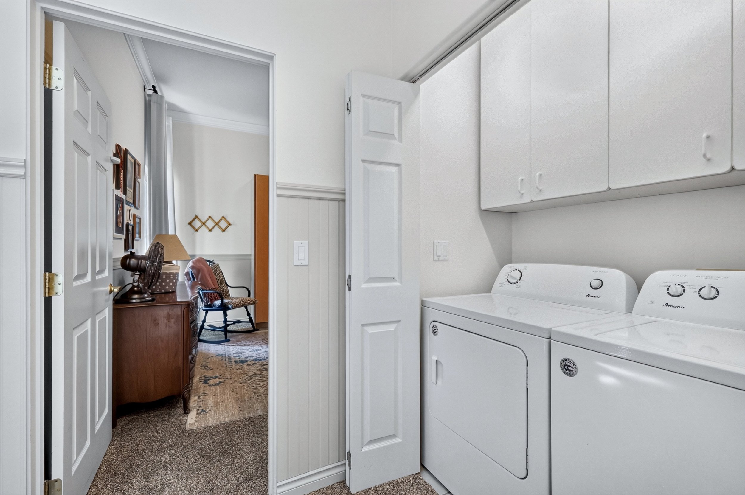 A laundry room with white washer and dryer units, white cabinets above, and a laundry room wall with hooks. An adjacent room in the background has a desk, chair, and framed pictures on the wall.