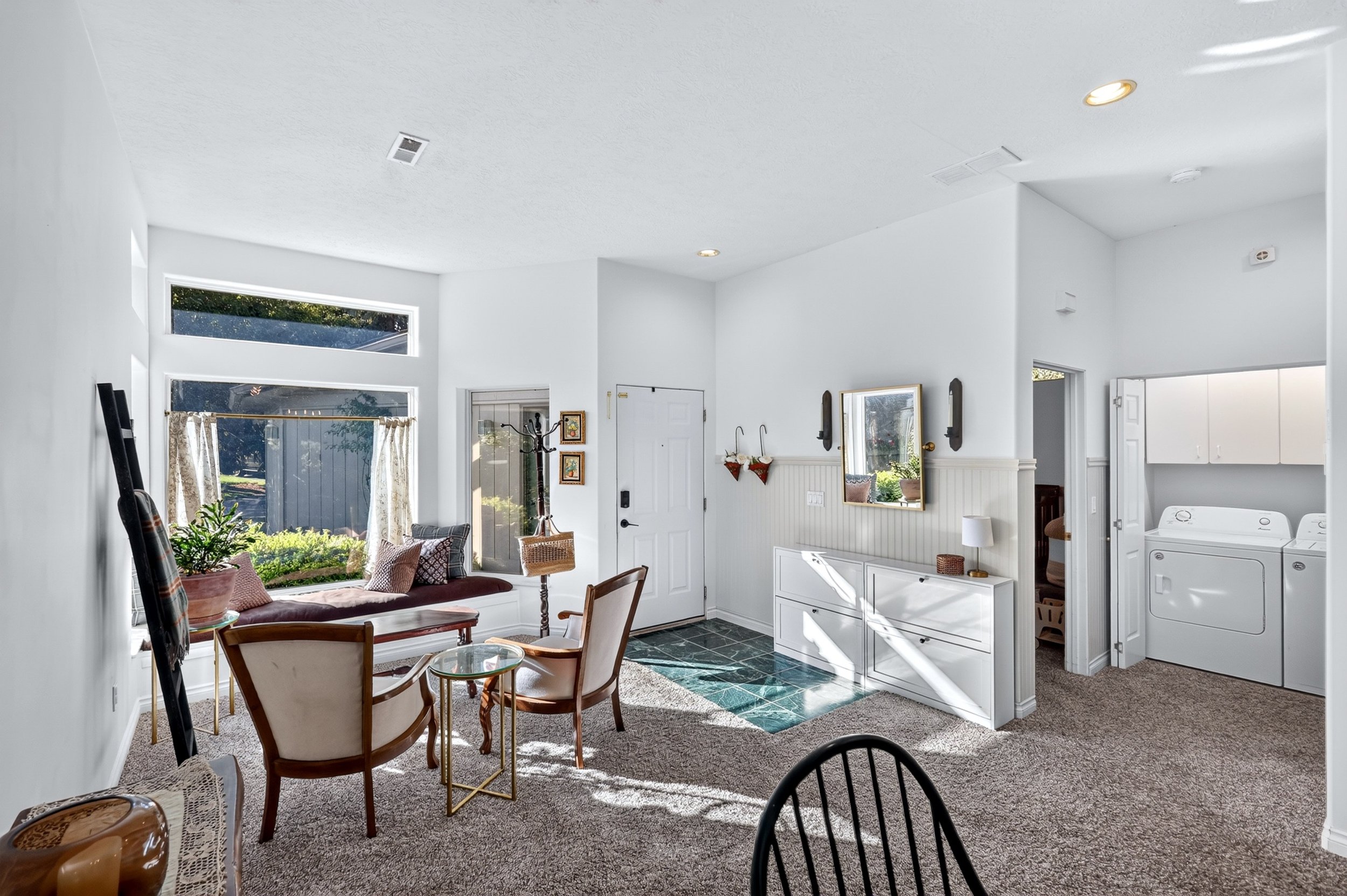 Bright, cozy living room with large front windows, a sitting area with a bench and chairs, green plants, and laundry appliances in the corner.