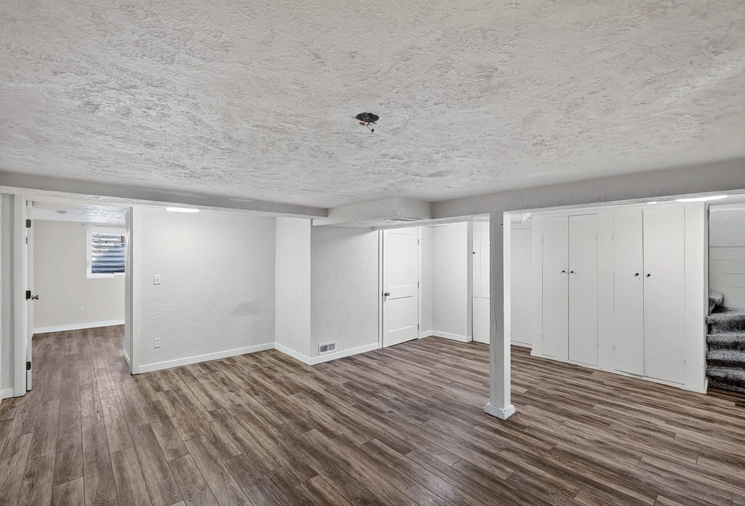 Empty room with white walls, wooden flooring, and a staircase carpeted in gray.

A section of ceiling has a small black object hanging from purple string, possibly a decorative item.