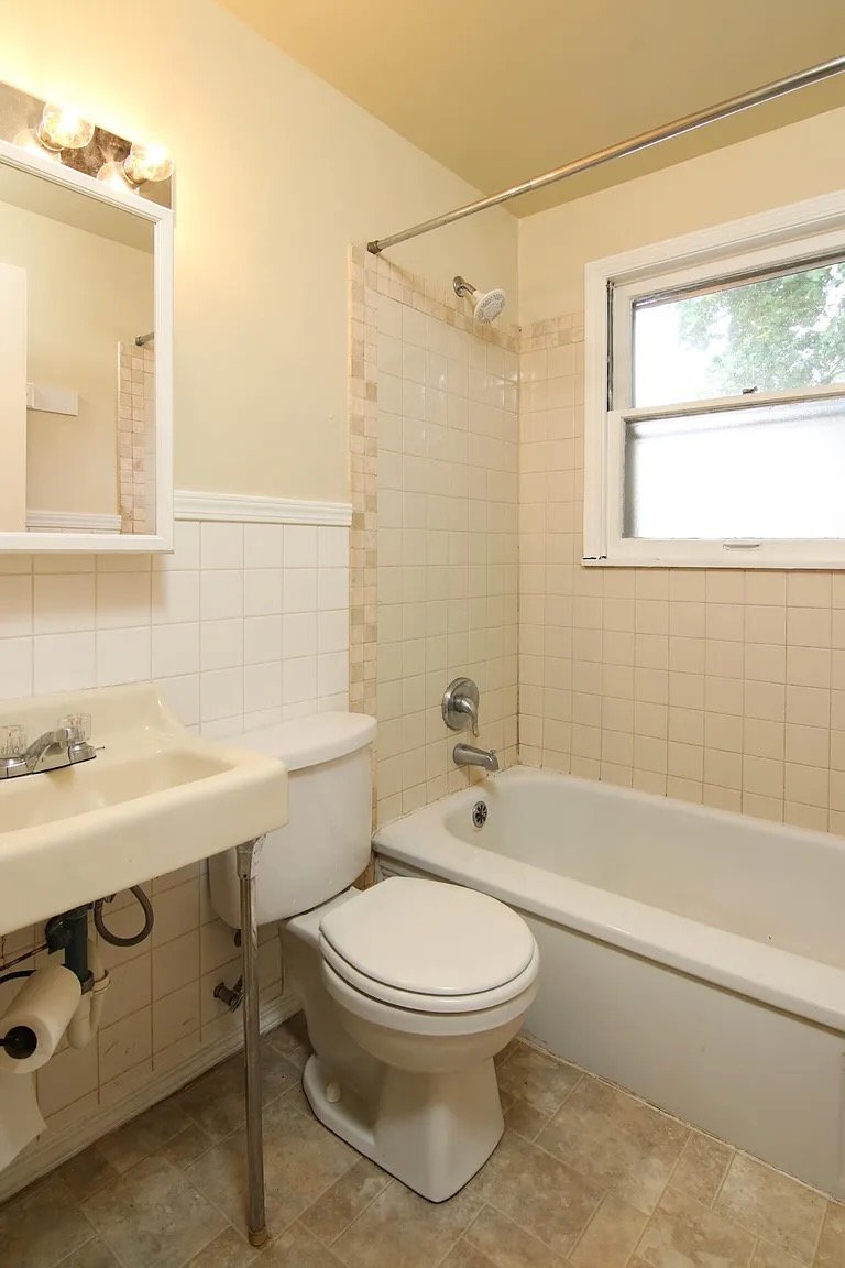 Small bathroom with a bathtub, toilet, small sink, window, and a mirror with light fixtures.