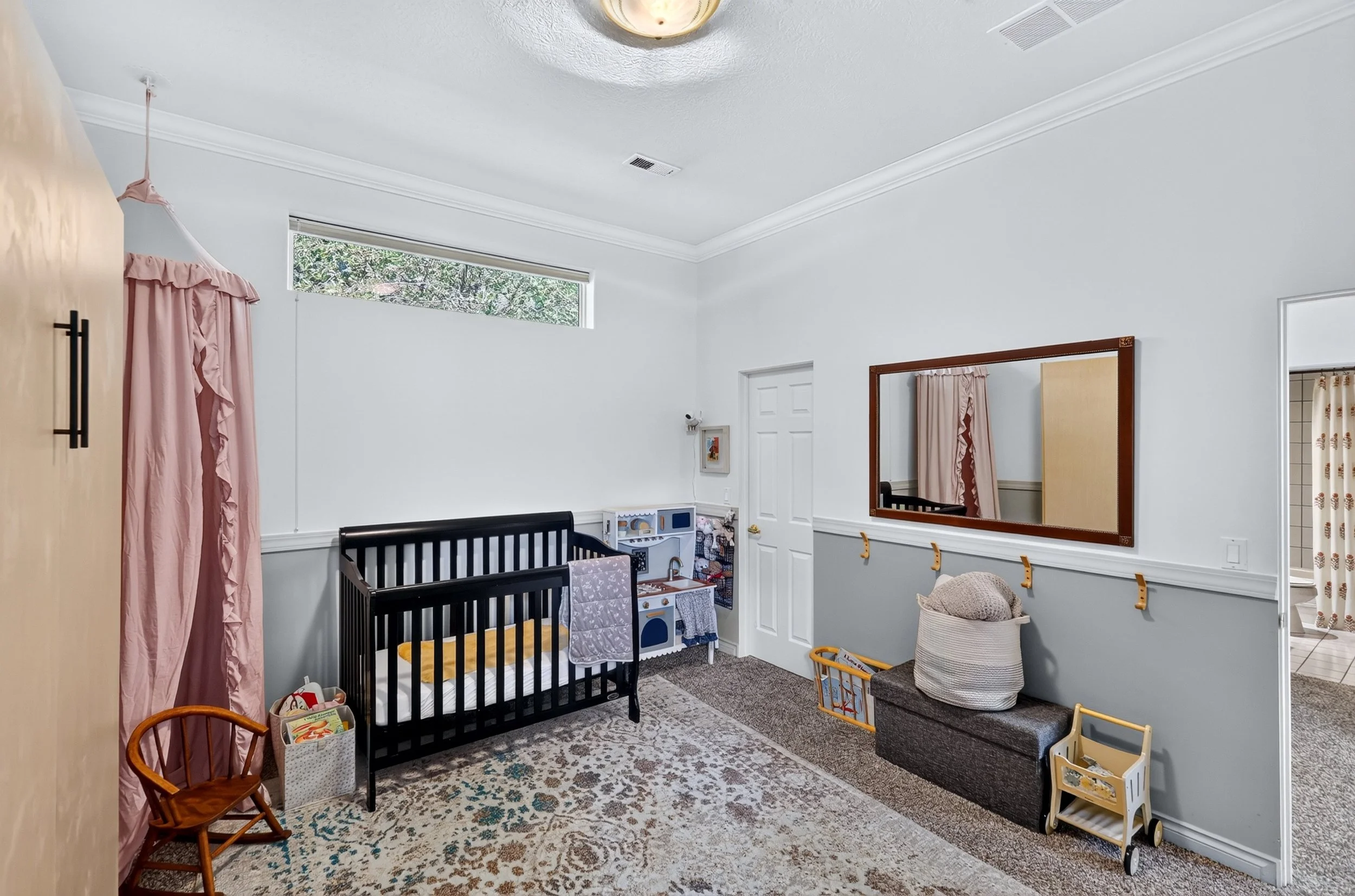 A nursery with a black crib, pink curtains, a white dollhouse, a small brown rocking chair, a basket, and a large mirror on the wall.