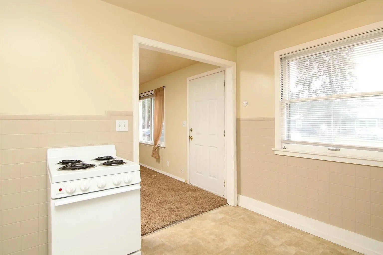 Empty kitchen with beige walls, large window with blinds, a white stove, and an entrance to another room with carpeted floor and a window with curtains.