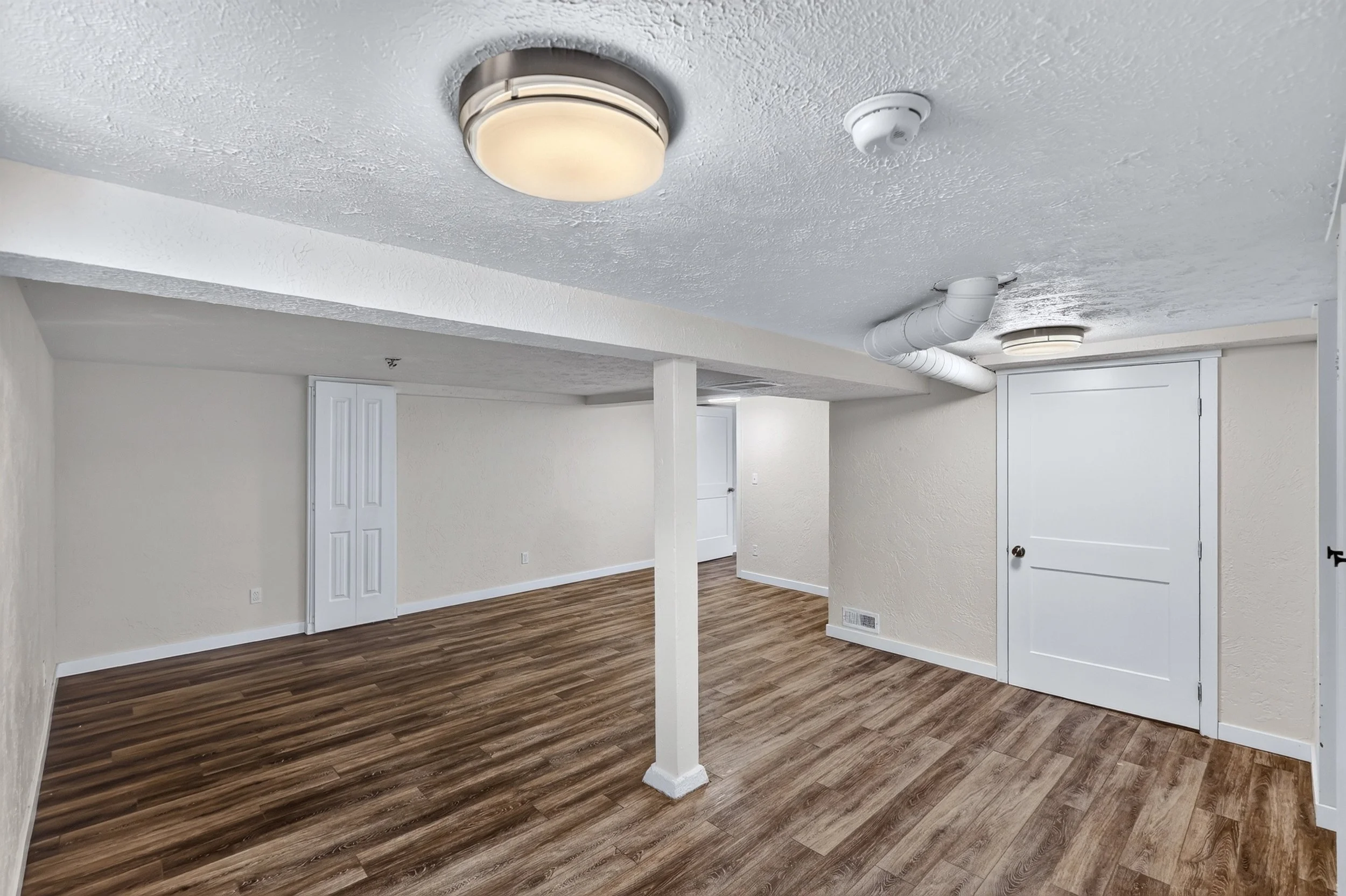 Empty room with textured white ceiling, white walls, wooden laminate flooring, ceiling light fixtures, smoke detector, HVAC vent, and a white door.