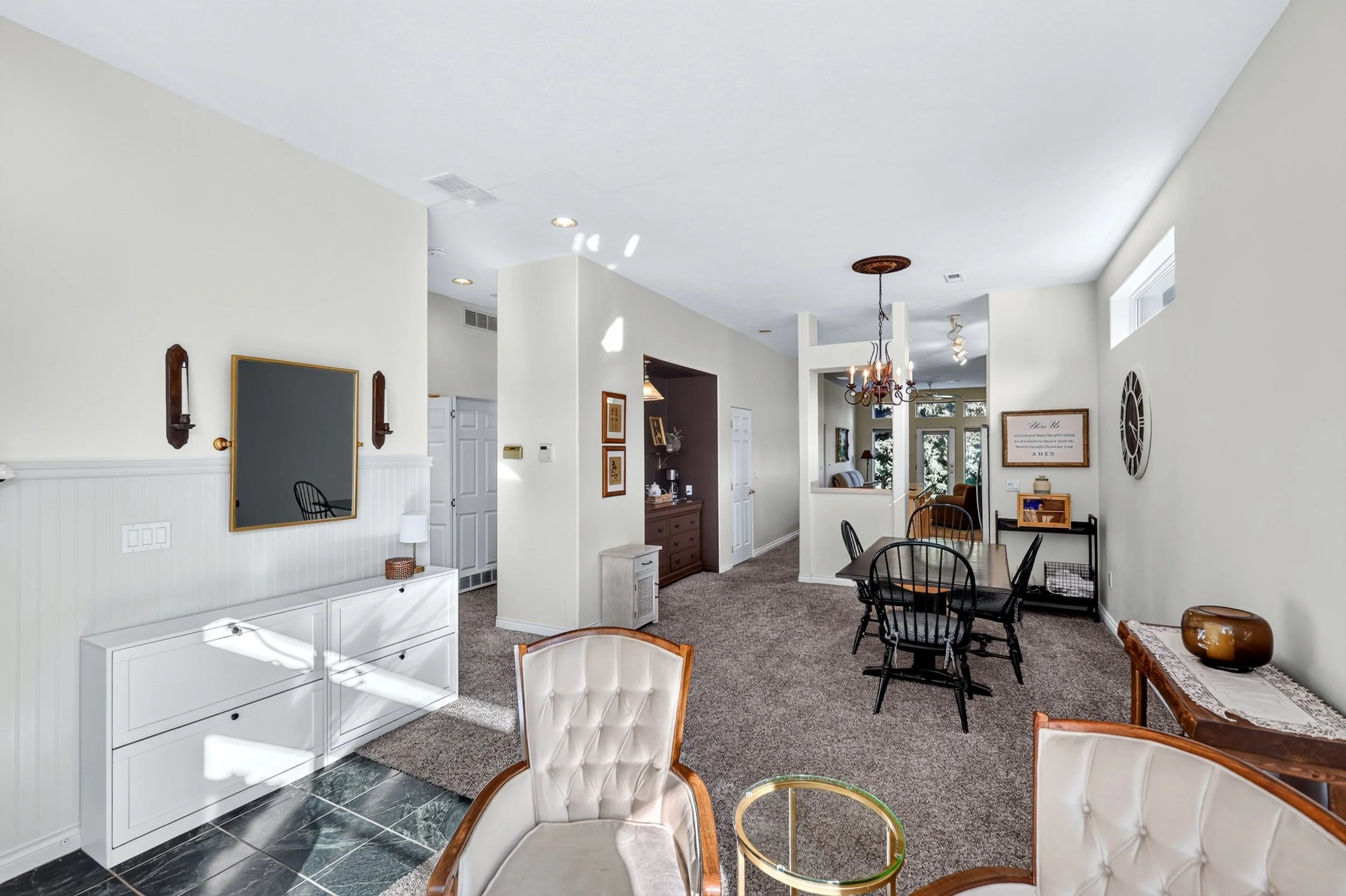 A spacious living and dining area with white walls, a chandelier, black dining chairs, artwork on the walls, and a window letting in natural light.
