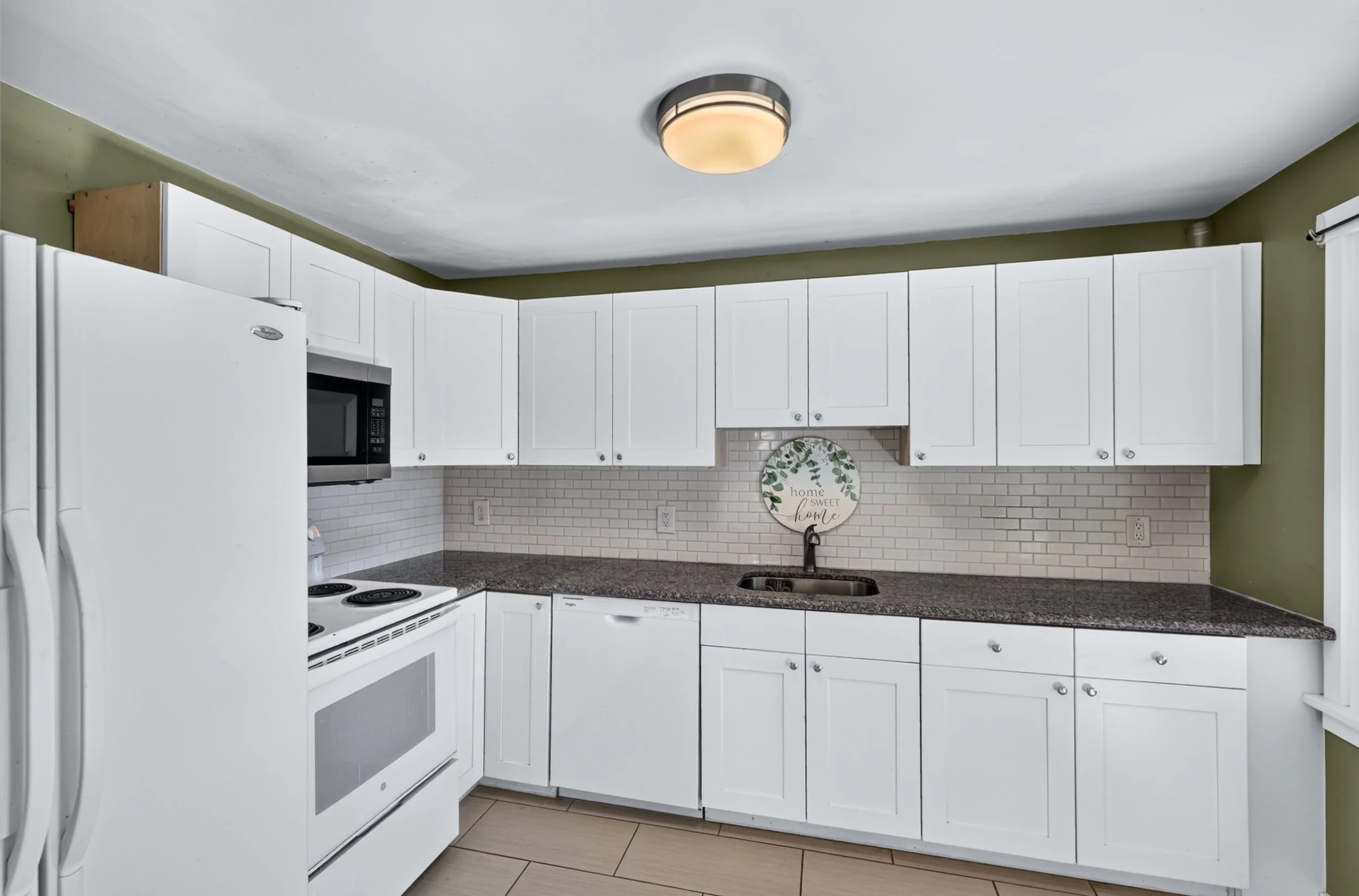White kitchen with white cabinets, black granite countertop, white stove, microwave, refrigerator, sink, tiled backsplash, and a decorative wall hanging that says 'home sweet home'