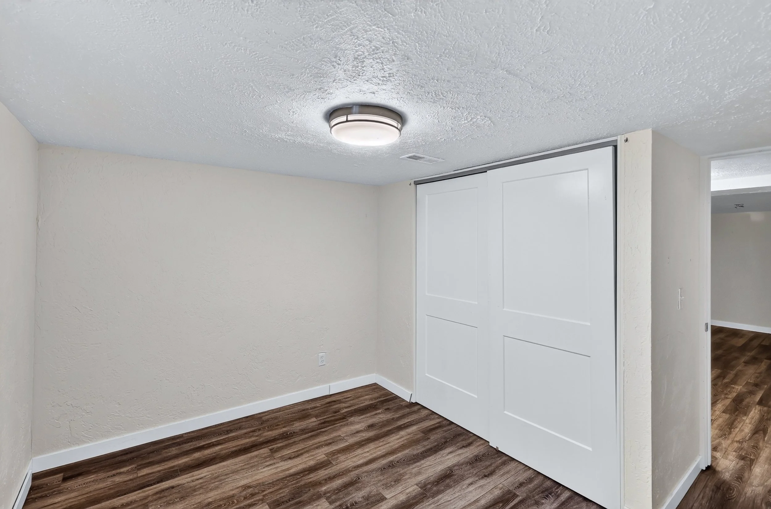 Interior of a room with white textured ceiling, beige walls, and brown wood flooring. There is a ceiling light fixture and a sliding white door.
