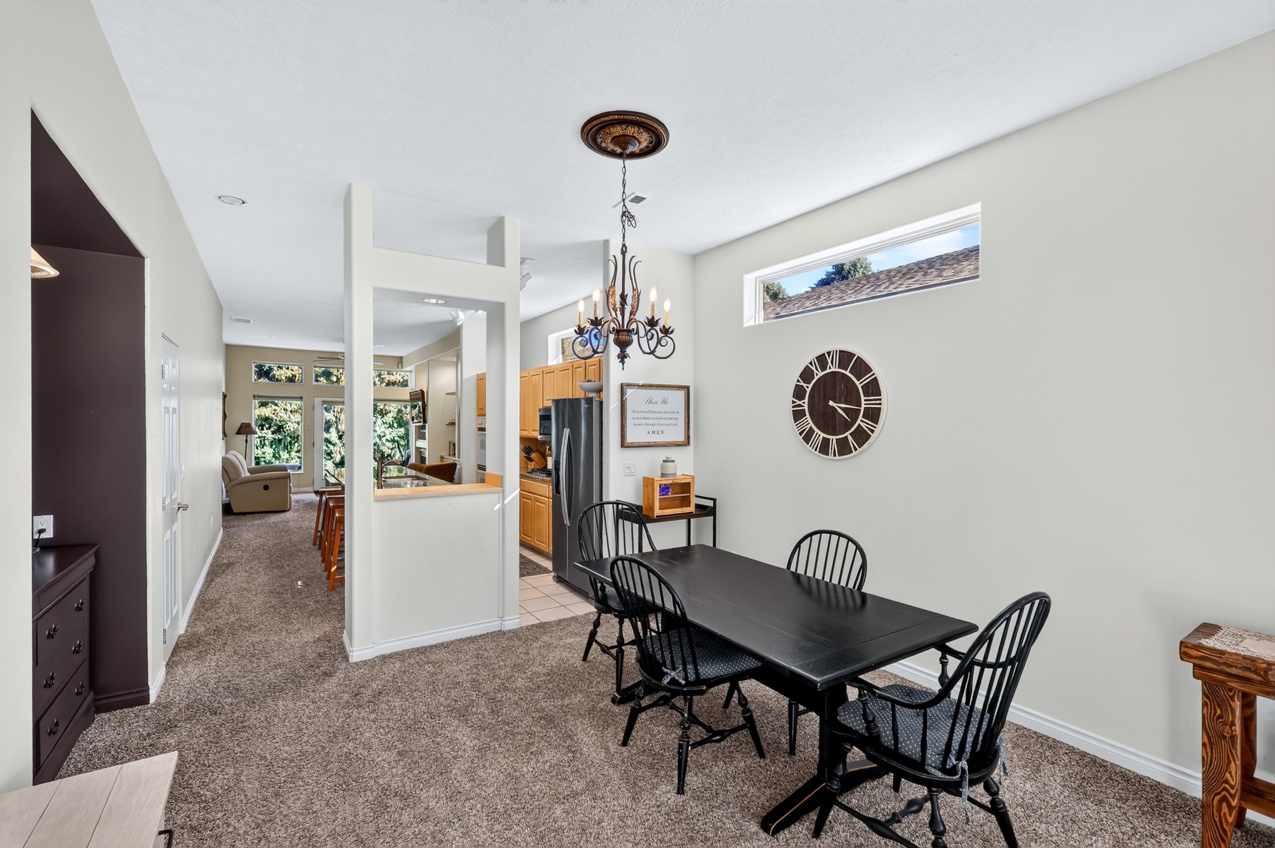 Interior view of a dining area and kitchen with white walls, a black dining table with matching chairs, a large clock on the wall, a chandelier, and a partial view of a living room with beige armchair and windows showing trees outside.