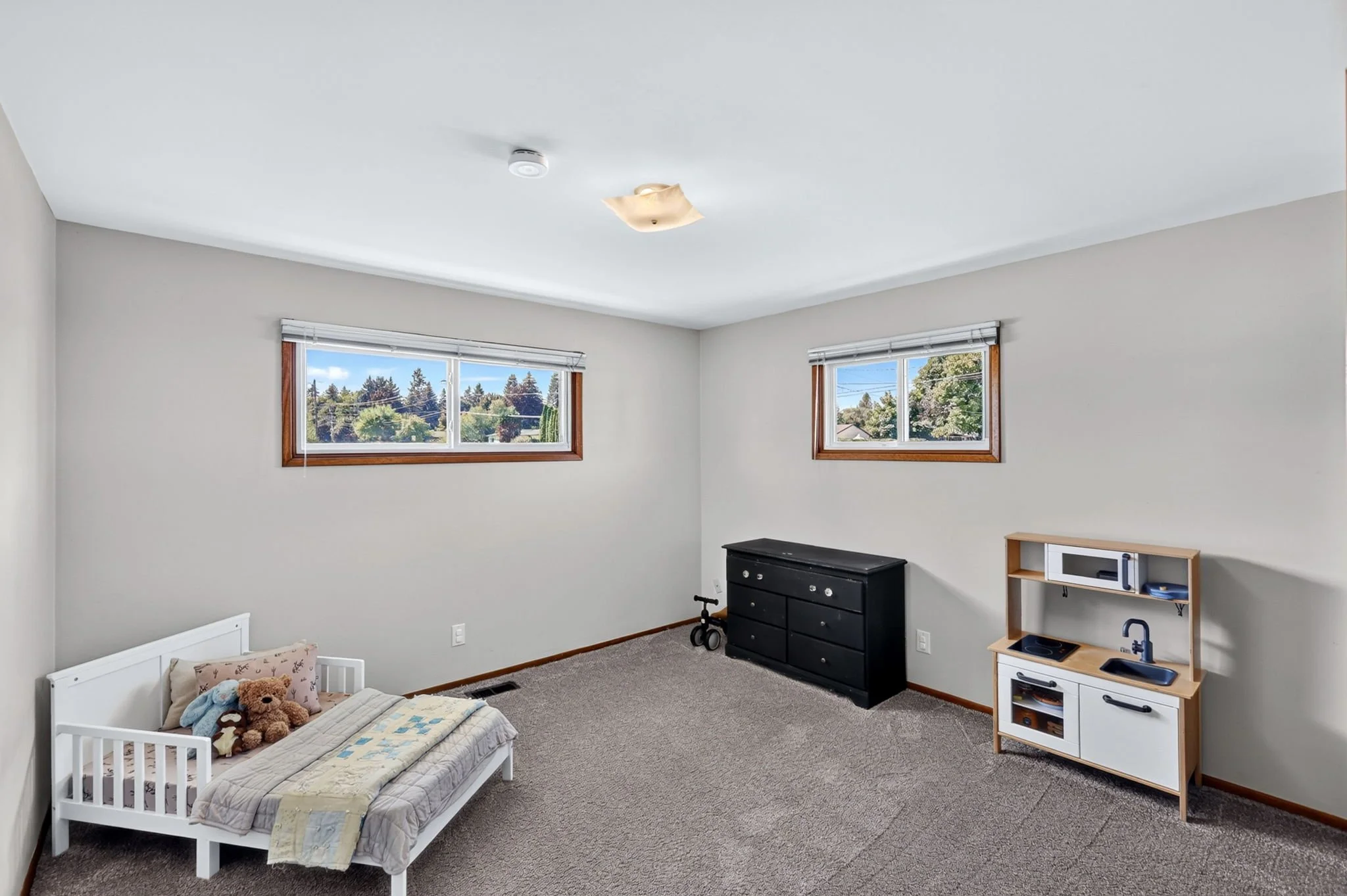 A bedroom with two windows, a small bed with stuffed animals, a black dresser, and a wooden play kitchen, beige carpet, and white walls.