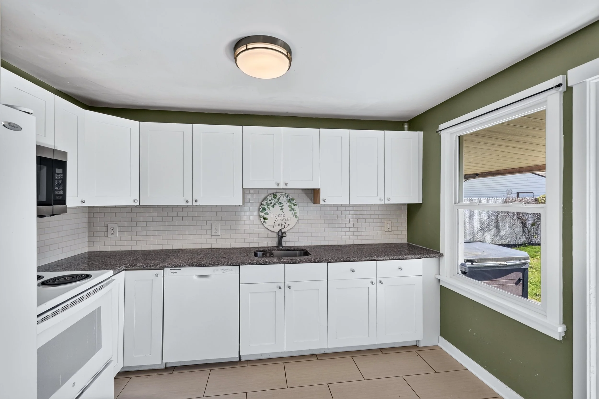 Kitchen with white cabinets, green walls, a window overlooking a backyard, white subway tile backsplash, and a round ceiling light fixture.