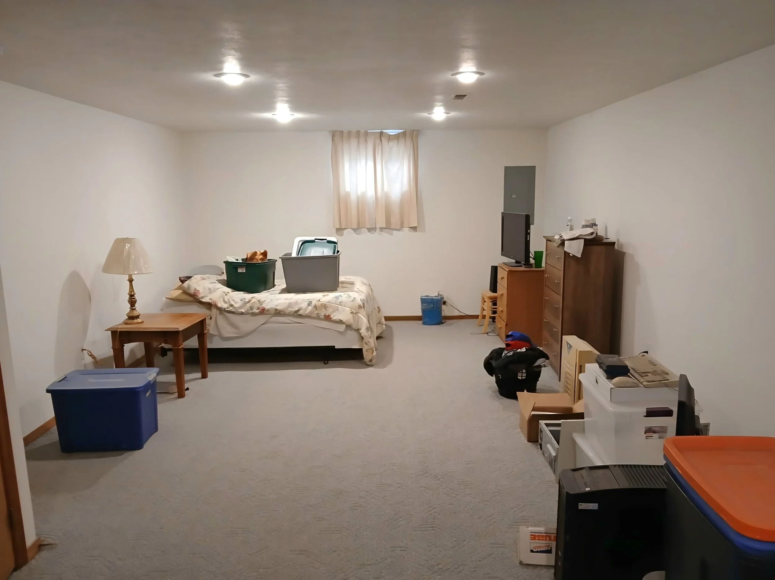 A cluttered living room with a bed pushed against the wall, a small wooden side table with a lamp on it, various storage bins and laundry baskets, a television on a wooden dresser, a window with beige curtains, and miscellaneous items scattered aroun
