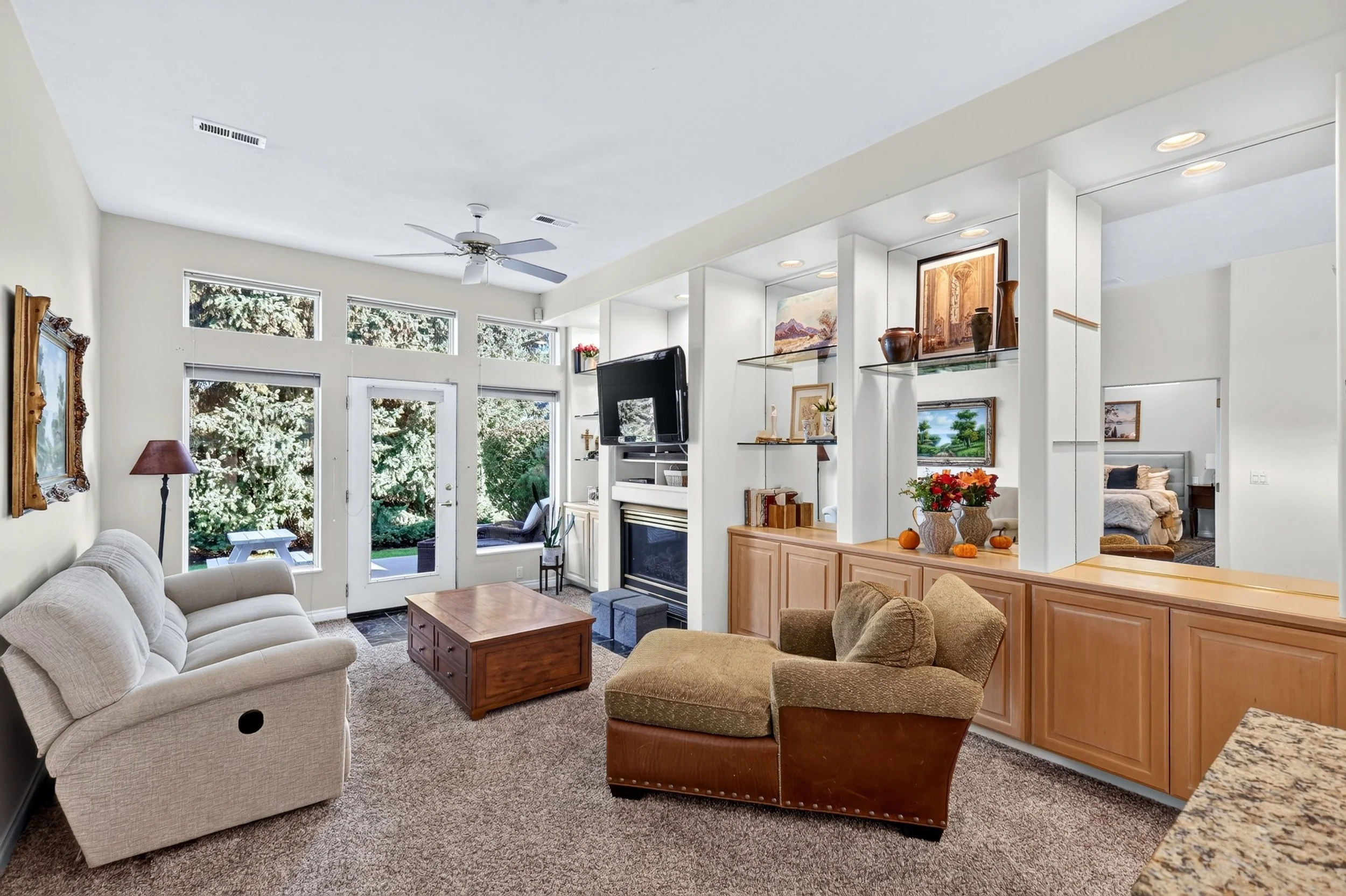 Living room with beige sofa and armchair, wooden coffee table, white walls, large windows, ceiling fan, built-in shelves with art and decor, fireplace, and view of a backyard with trees