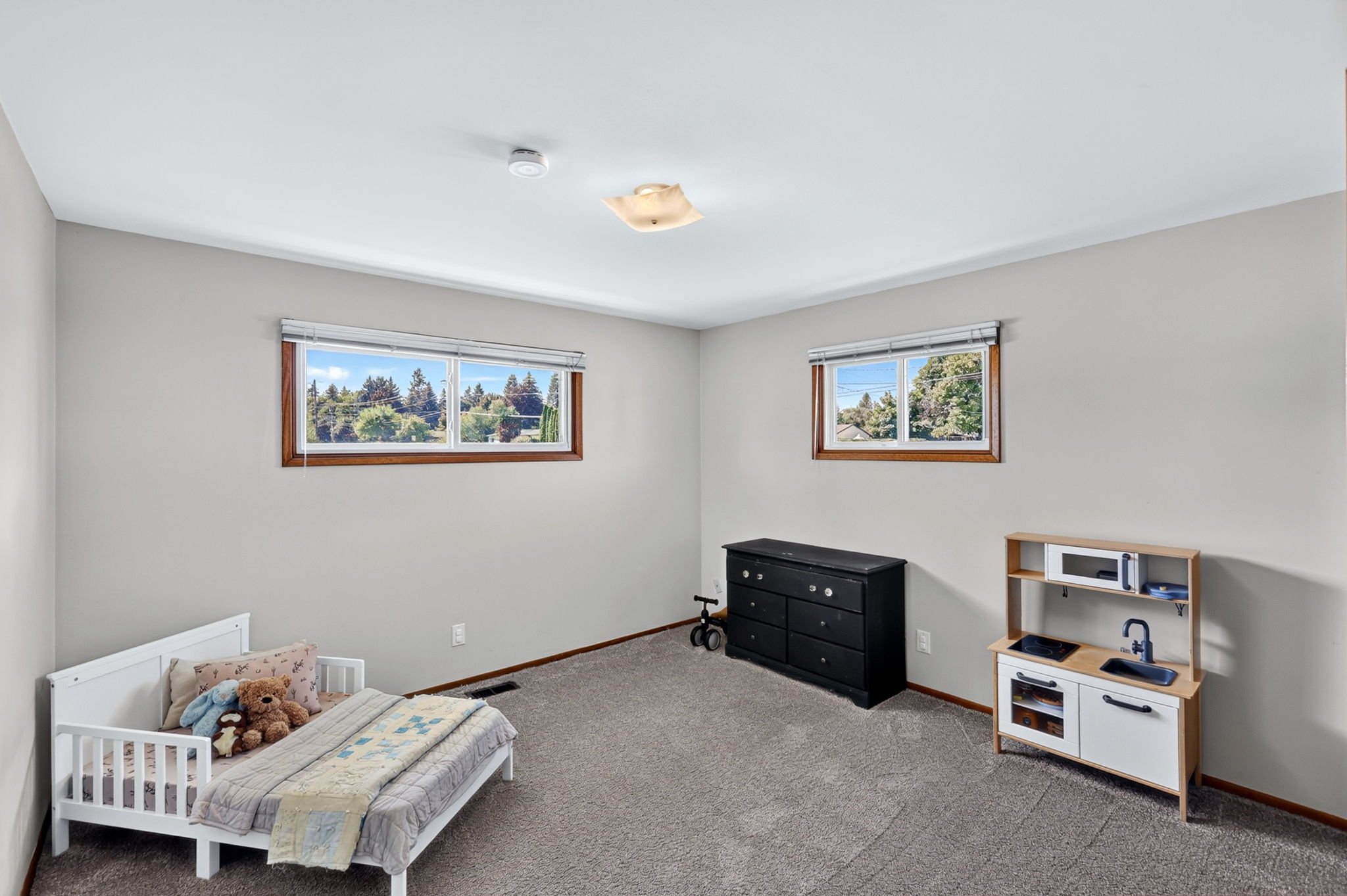 Empty child's bedroom with small bed, stuffed animals, black dresser, and toy kitchen, two windows with views of trees outside, beige walls, carpeted floor.