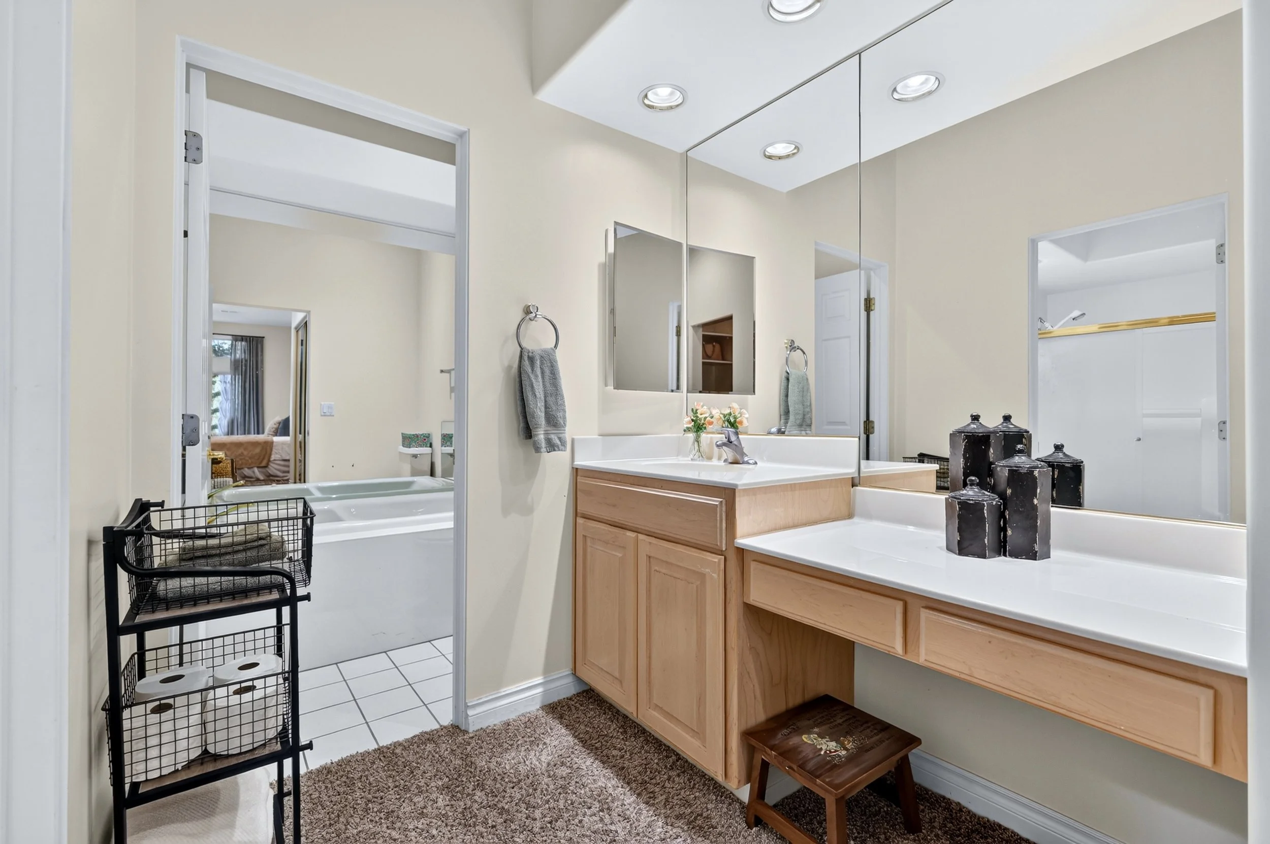 A bathroom with a double sink vanity, frameless mirror, and recessed ceiling lights. There are towels hanging on rings and decorative candles on the countertop. A small wooden step stool is in front of the vanity, and a black metal storage rack with 
