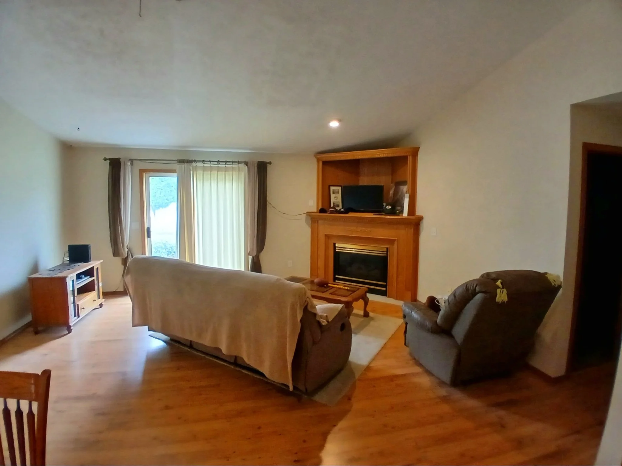 Living room with beige walls, a wooden fireplace, two sofas, a TV on a wooden stand, a window with curtains, and hardwood flooring.