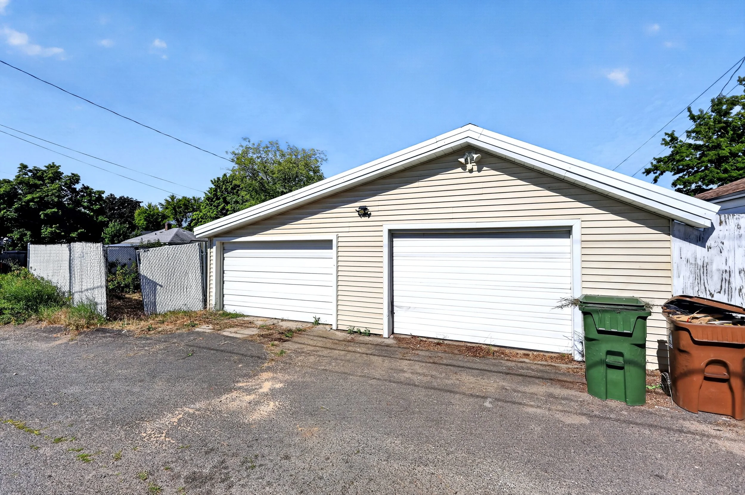 The house with beige siding, two white garage doors, security cameras, and outdoor lights, with trash bins outside, is surrounded by trees and a chain-link fence, under a clear blue sky.