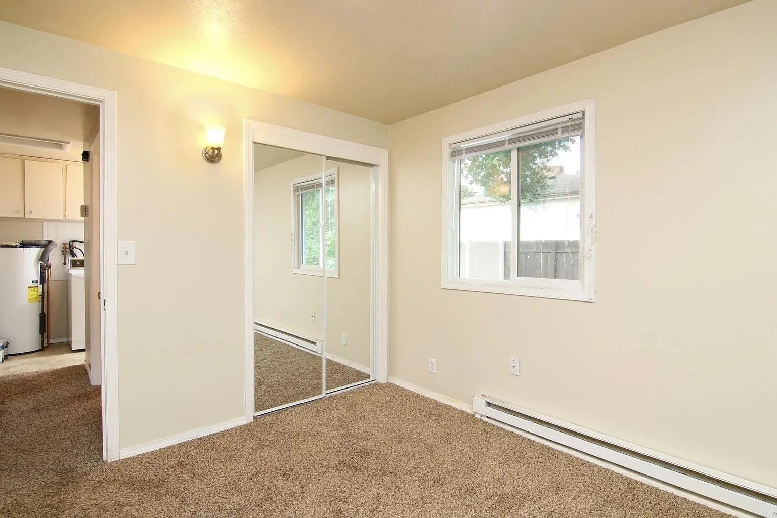 Empty room with beige walls, brown carpet, a window with a view of a backyard, a closet with mirrored sliding doors, a wall-mounted light fixture, and baseboard heating.