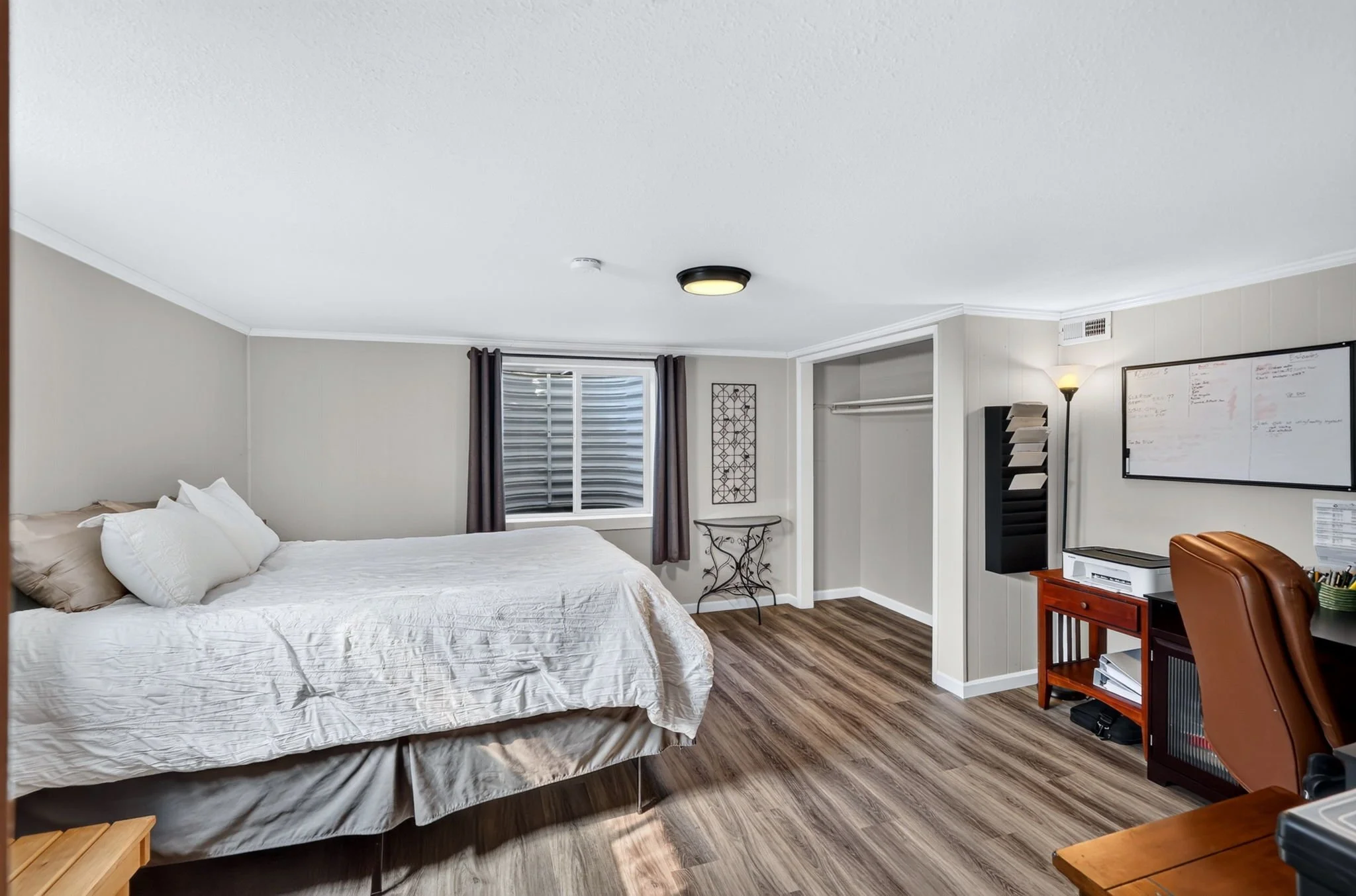 A bedroom with a large bed covered with a white quilt, gray pillows, and a small wooden side table. There is a window with dark curtains, a decorative wall art piece, a small round table, a whiteboard on the wall, a black filing cabinet, a desk with 